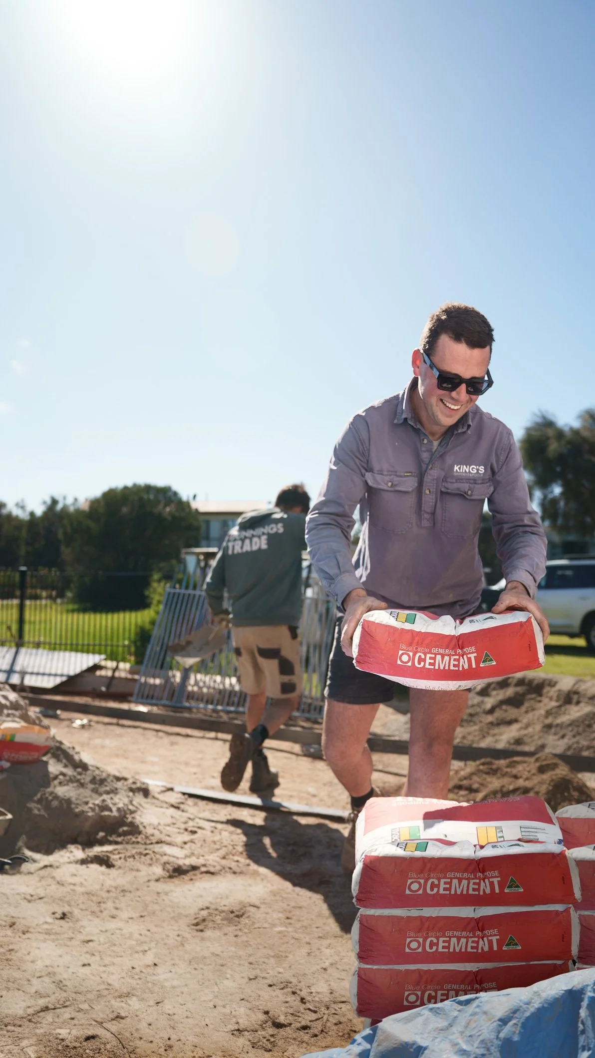 Two men working outdoors on a sunny day, one smiling and stacking cement bags, the other carrying a cement bag in the background.