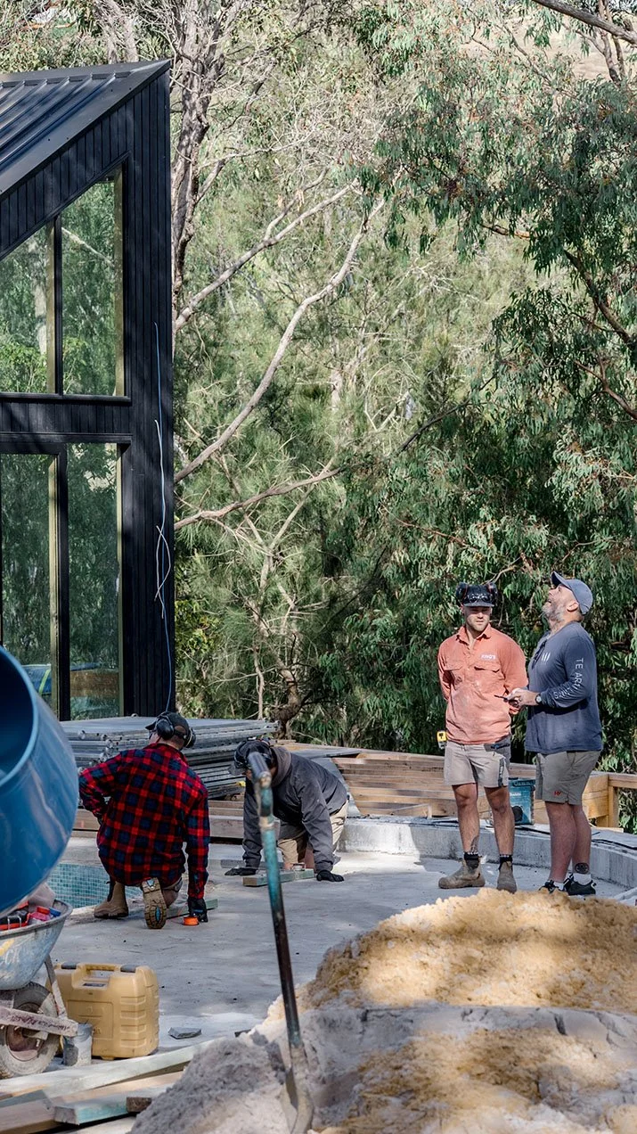 Construction workers building a deck or patio outdoors next to a modern black house with large glass windows, surrounded by trees.