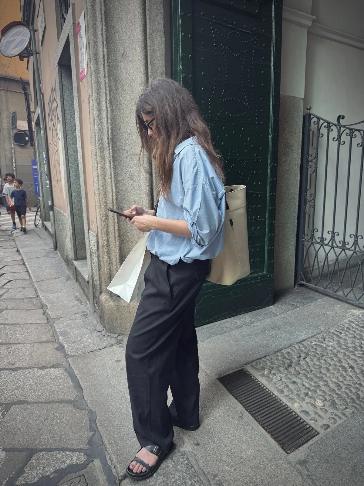 A woman with long brown hair wearing glasses, a light blue shirt, black pants, and black sandals, looking at her phone while standing on a city sidewalk next to a green iron gate.