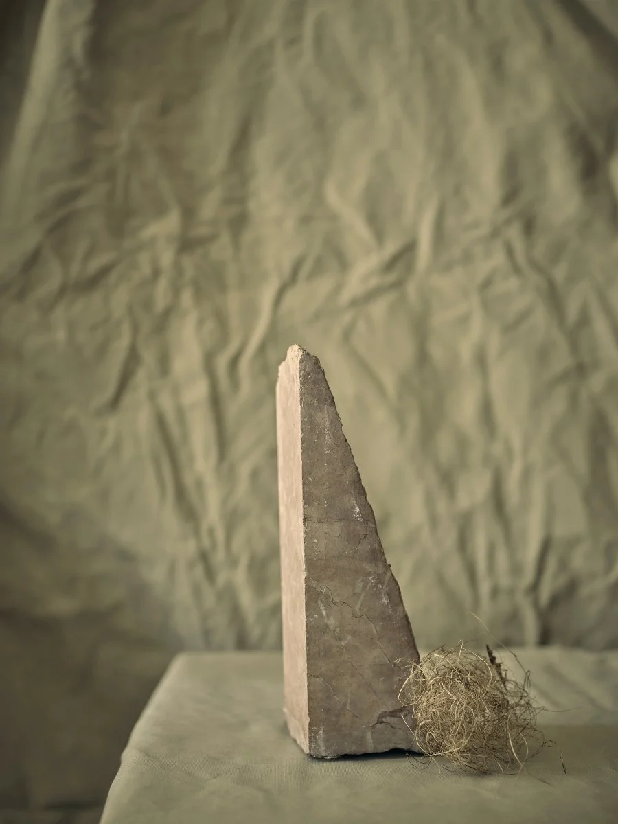A triangular concrete or stone object with a rough edge, resting on a flat surface with a small bundle of frayed twine or string beside it, against a textured background.