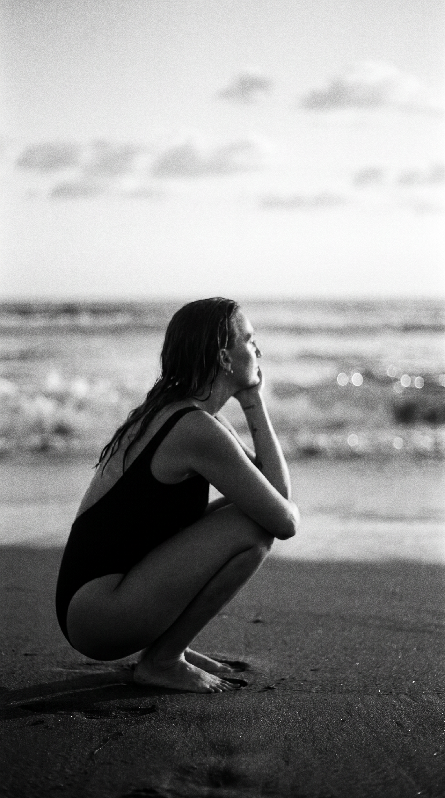 A person crouches on the beach in a black swimsuit, staring at the ocean, with wet hair, in a contemplative pose, in black and white.