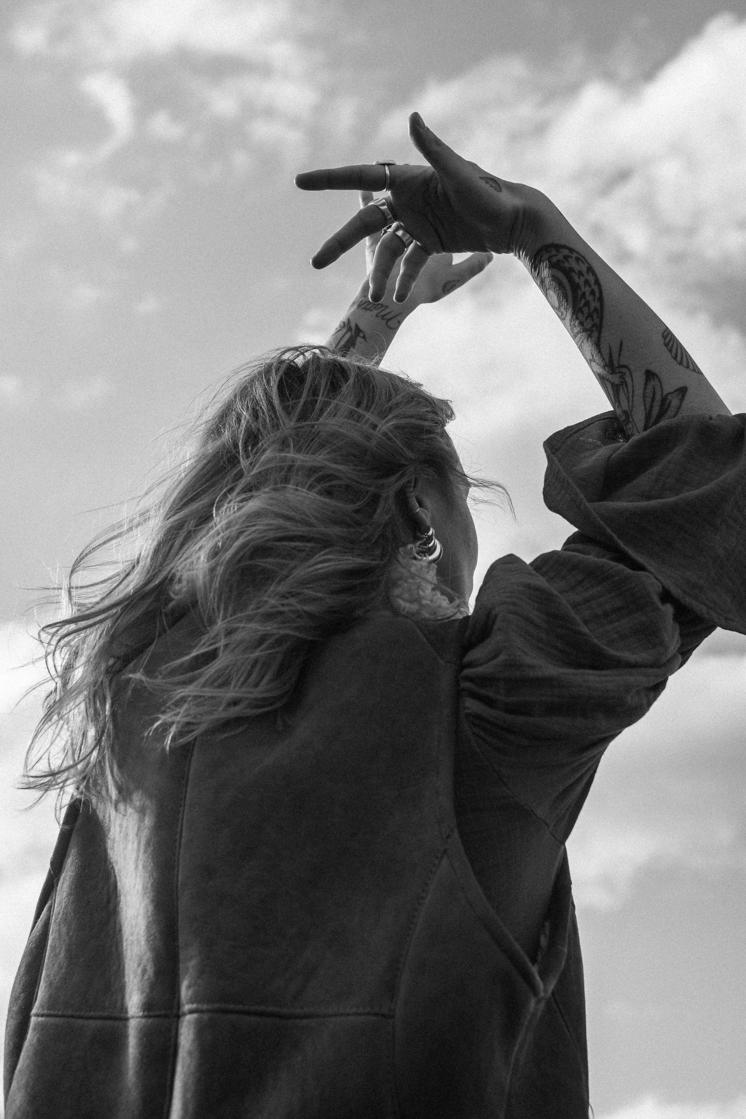 A person with tattoos on their arms raising their hands in the air with clouded sky in the background.