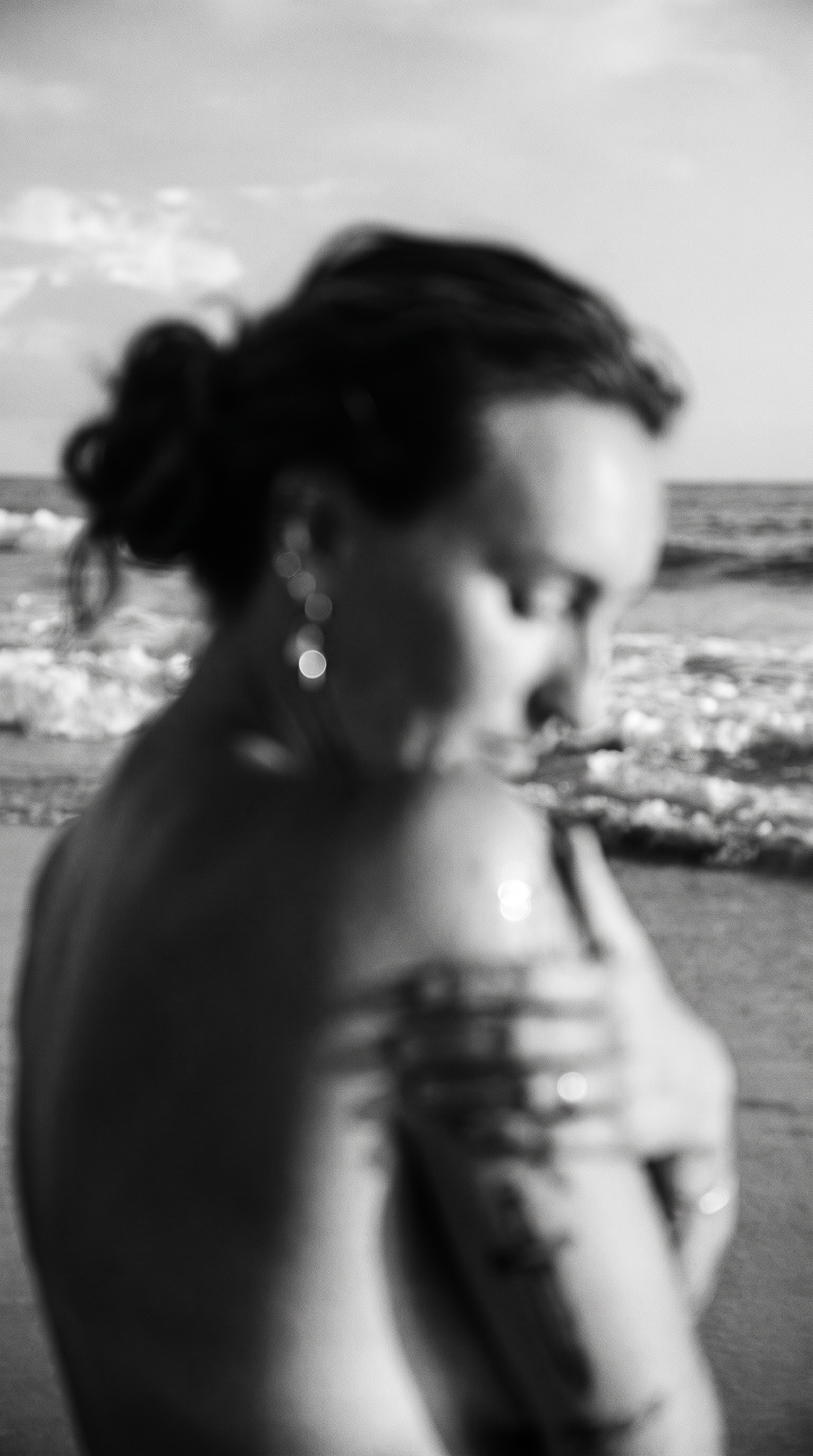 A woman with short dark hair and earrings stands on a beach, looking down at her arm, while holding her shoulder with her hand.