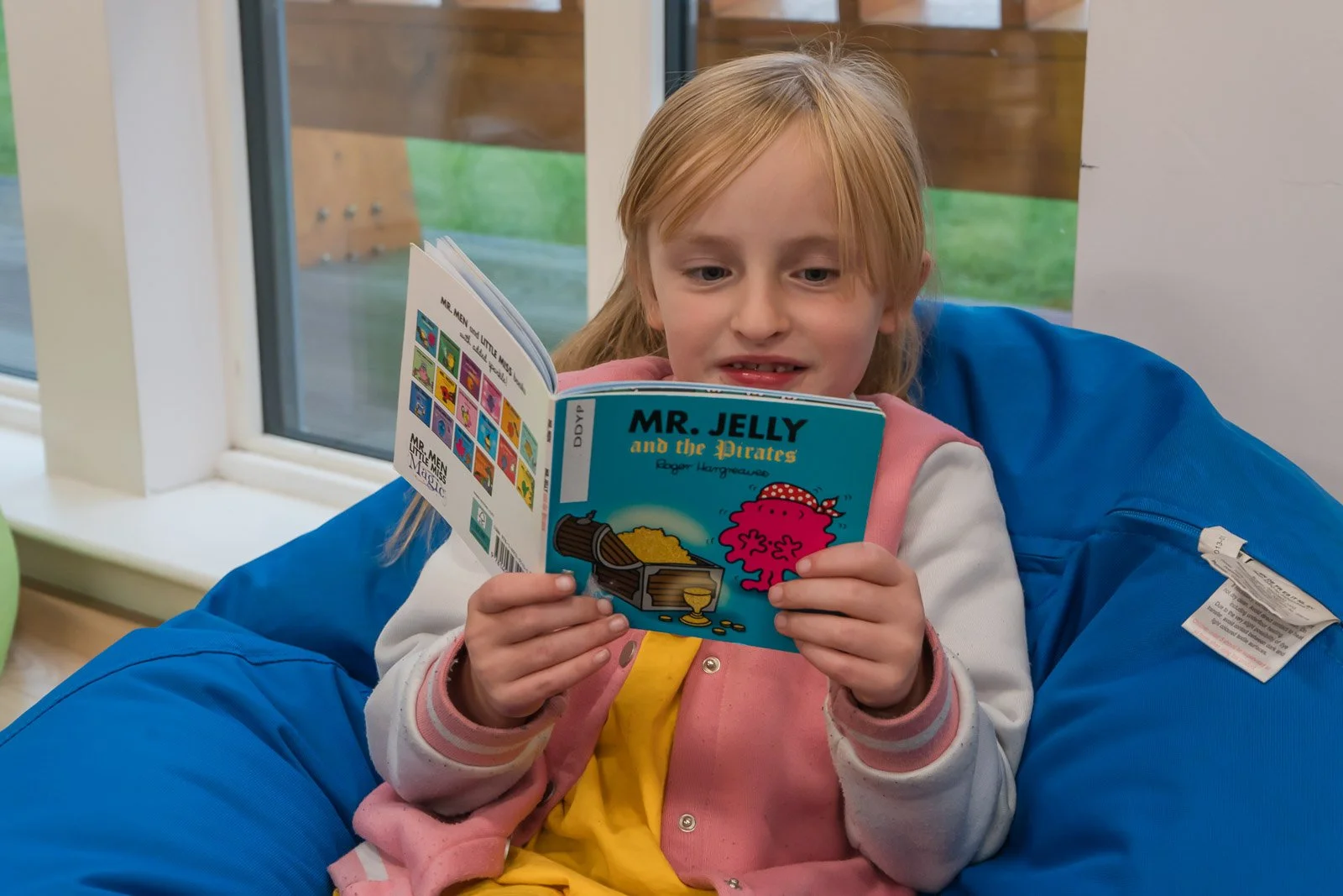 A young girl with red hair is sitting indoors, wearing a pink jacket over a yellow shirt, reading a colourful comic book titled 'Mr. Jelly and the Pirates' while sitting on a large blue bean bag chair near a window. DDYP Doune