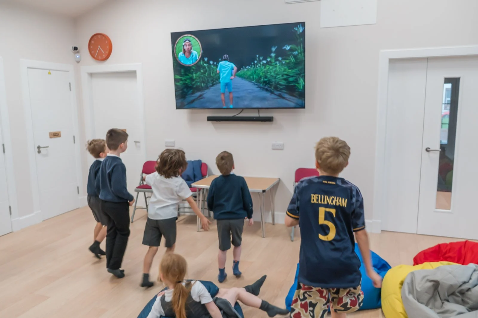 A group of children watching a virtual reality experience on a large television screen in a classroom or activity room. The screen shows a person in a blue shirt running in a digital outdoor setting. This is the after schools club in Doune - DDYP 