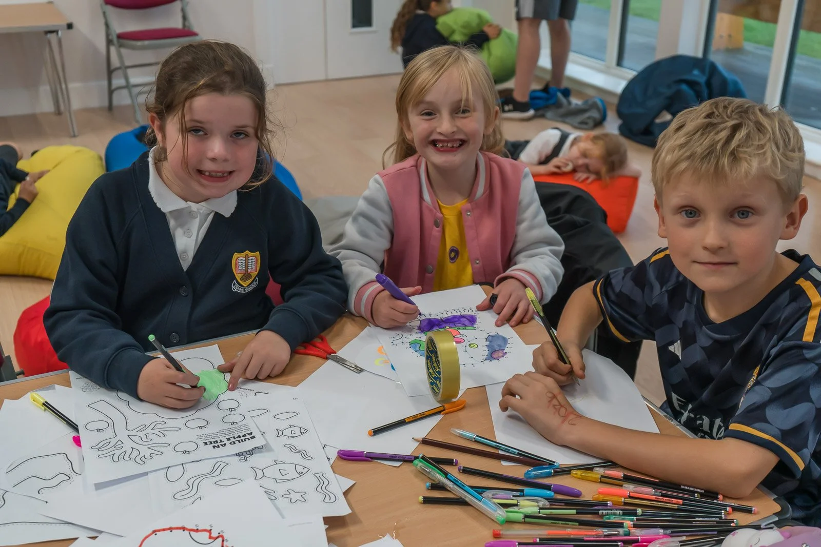 Three children are sitting at a table engaged in colouring activities, with colouring pages, markers, and colouring supplies scattered on the table, and a girl in the background near a large window in the DDYP pavilion. 