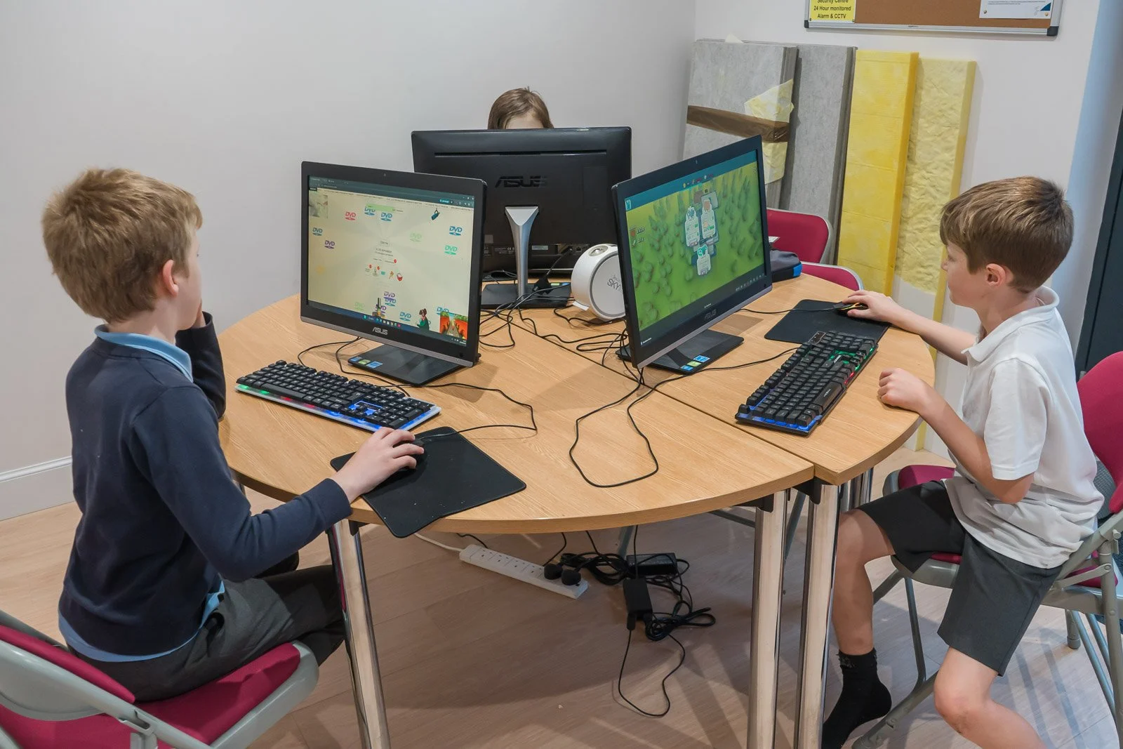 Two boys are sitting at a round table using desktop computers, with a woman in the background working at a computer. The boys are focused on their screens. This is the after-school club at DDYP in Doune.