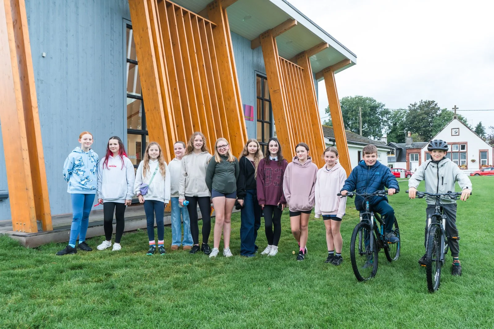 A group of twelve children, mostly girls and two boys, standing in a line on a grassy area in front of of the DDYP pavillion.