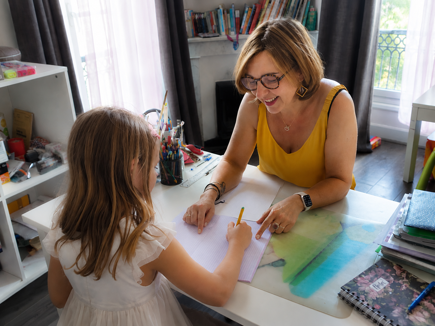 Une femme et une jeune fille travaillent ensemble sur une tâche scolaire ou créative à une table à la maison, avec des fournitures colorées autour d'eux.