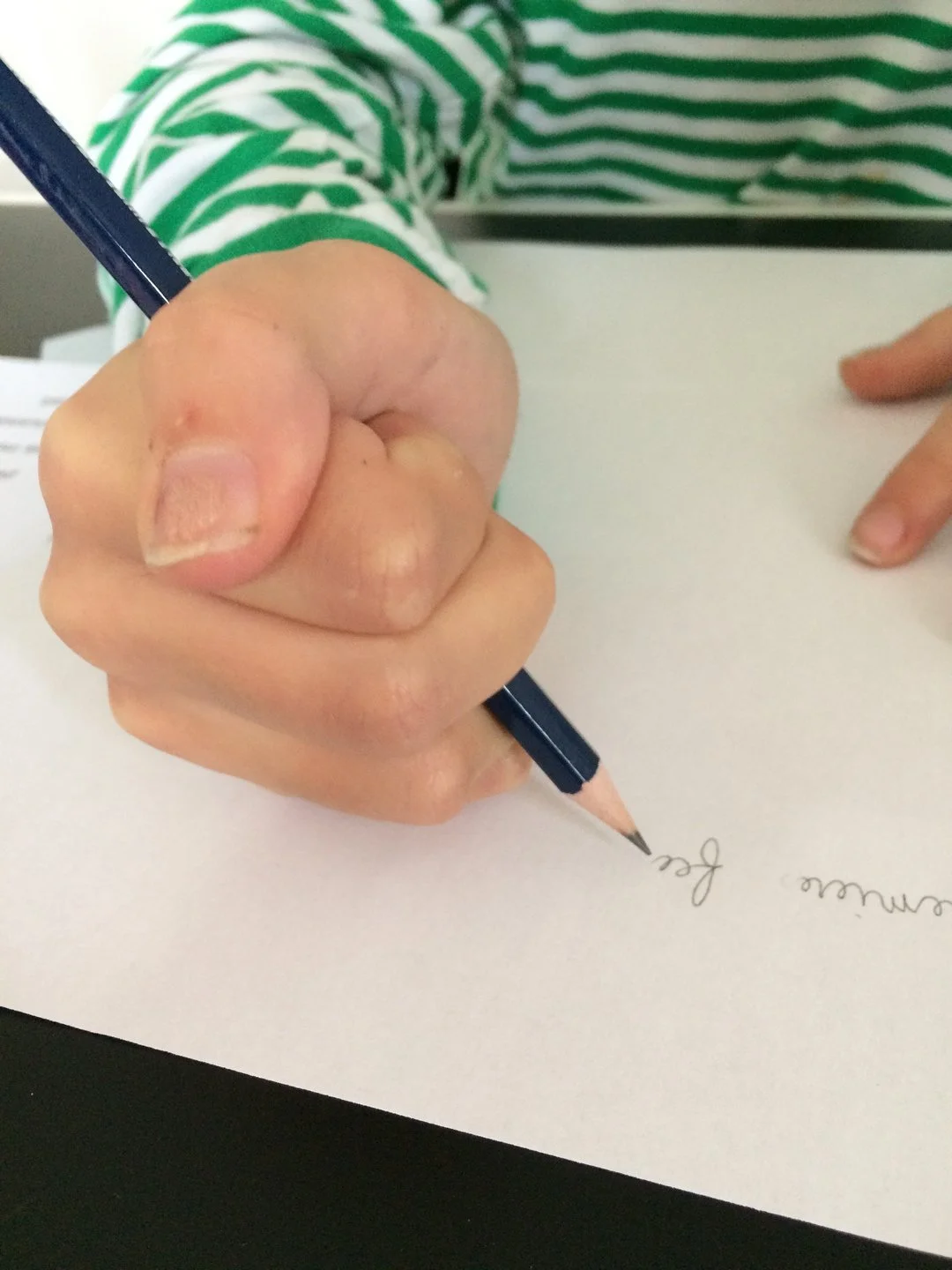 Une main qui écrit au crayon sur une feuille blanche, portant un vêtement rayé vert et blanc.