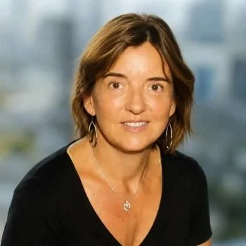 Portrait d'une femme avec des cheveux bruns, portant des boucles d'oreilles et un collier, fond flou de ville