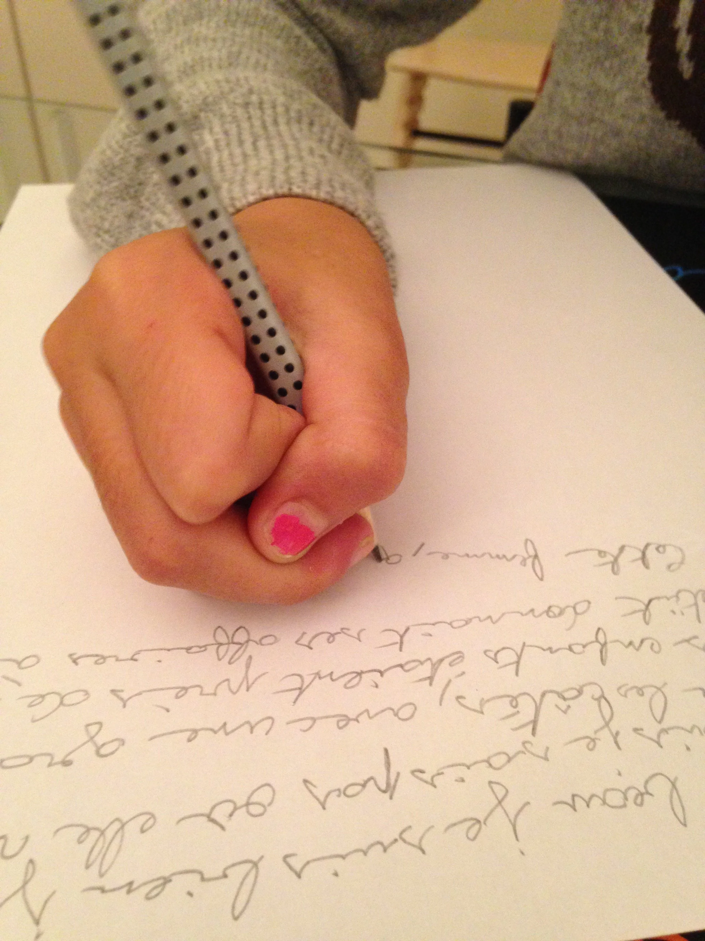Main tenant un stylo gris à pois noirs, écrivant sur une lettre manuscrite avec du texte en cursive, posé sur une table. Le fond montre une personne en pull gris clair et un meuble en bois.