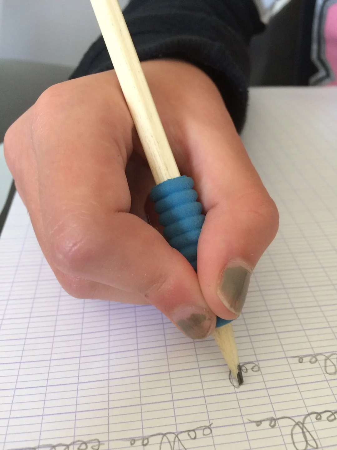 Une main tenant un stylo en bois avec une poignée bleue, écrivant sur une feuille de papier quadrillée. La main a des ongles peints en gris.
