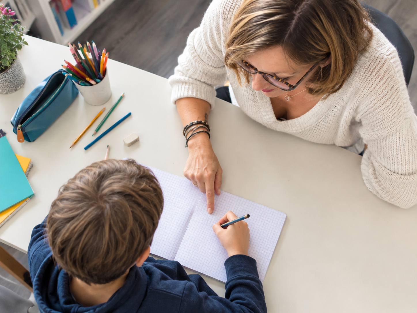 Une femme enseignant une jeune garçon à écrire dans un cahier à carreaux sur une table blanche, avec des crayons, un stylo et des fournitures scolaires autour.