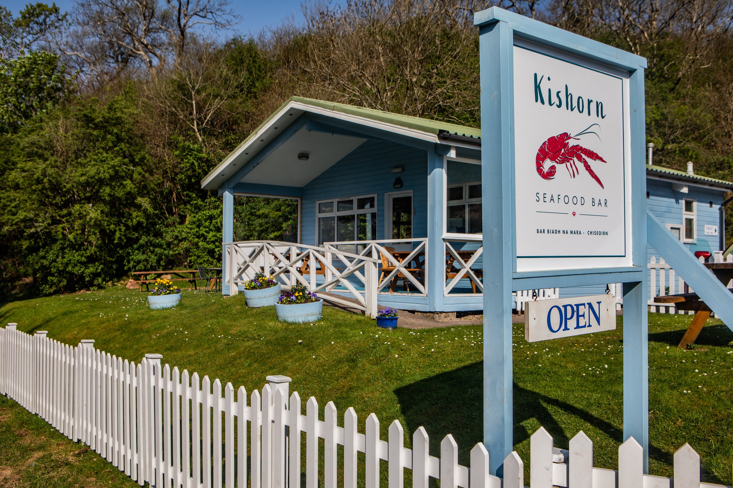 Blue building with a sign that says 'Kishorn Seafood Bar' featuring a red lobster illustration, surrounded by a white picket fence, potted flowers, and a lush green background.