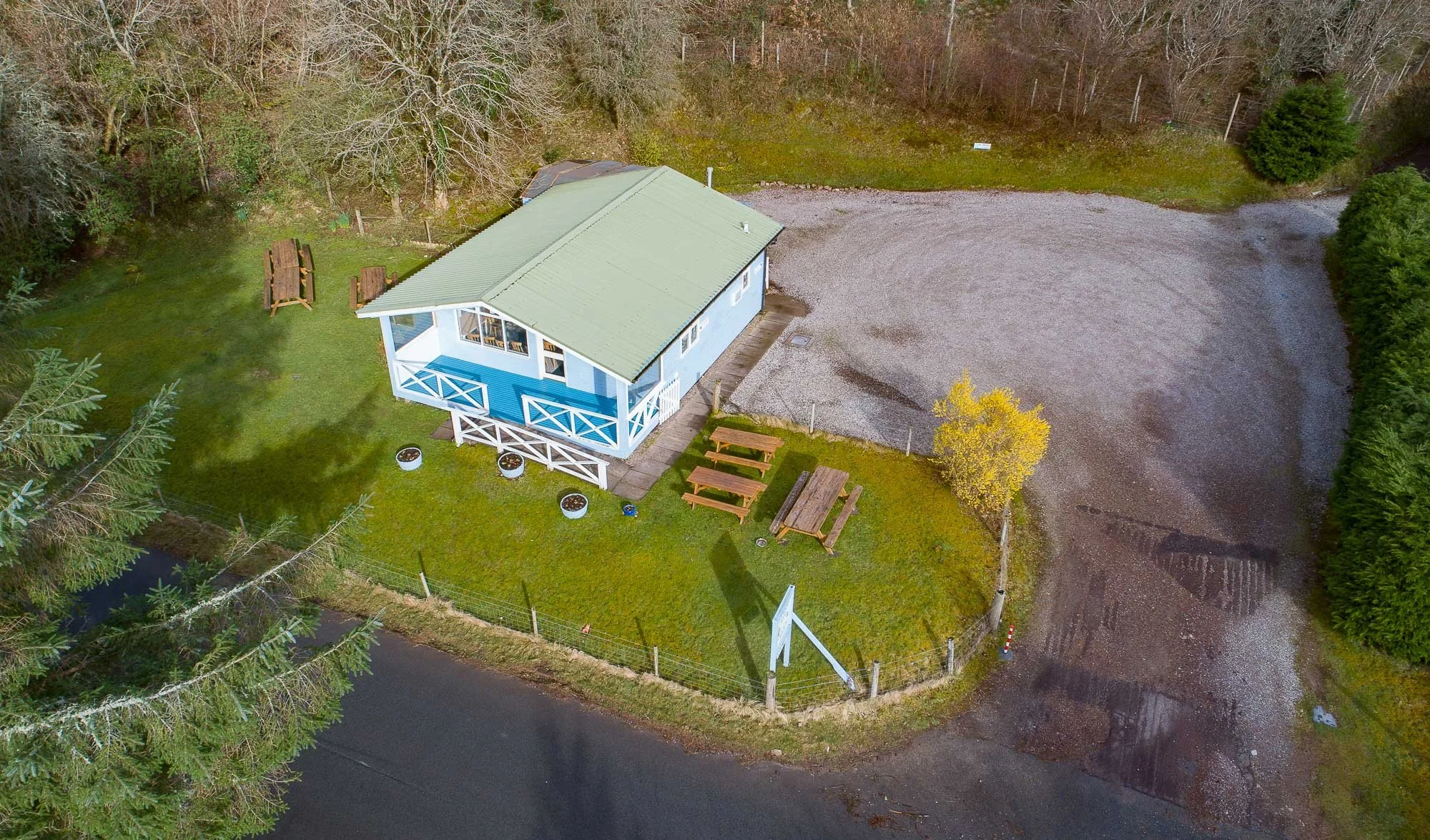 Aerial view of Kishorn Seafood Bar, surrounded by a lawn with picnic tables, trees, a gravel driveway, and a fenced yard.