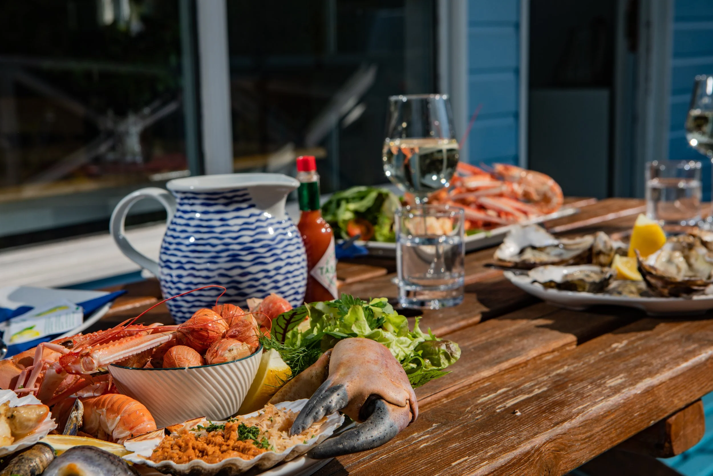 Seafood dinner on a wooden table with oysters, lobster, shrimp, fish, and various side dishes, set near a window with a house exterior visible.