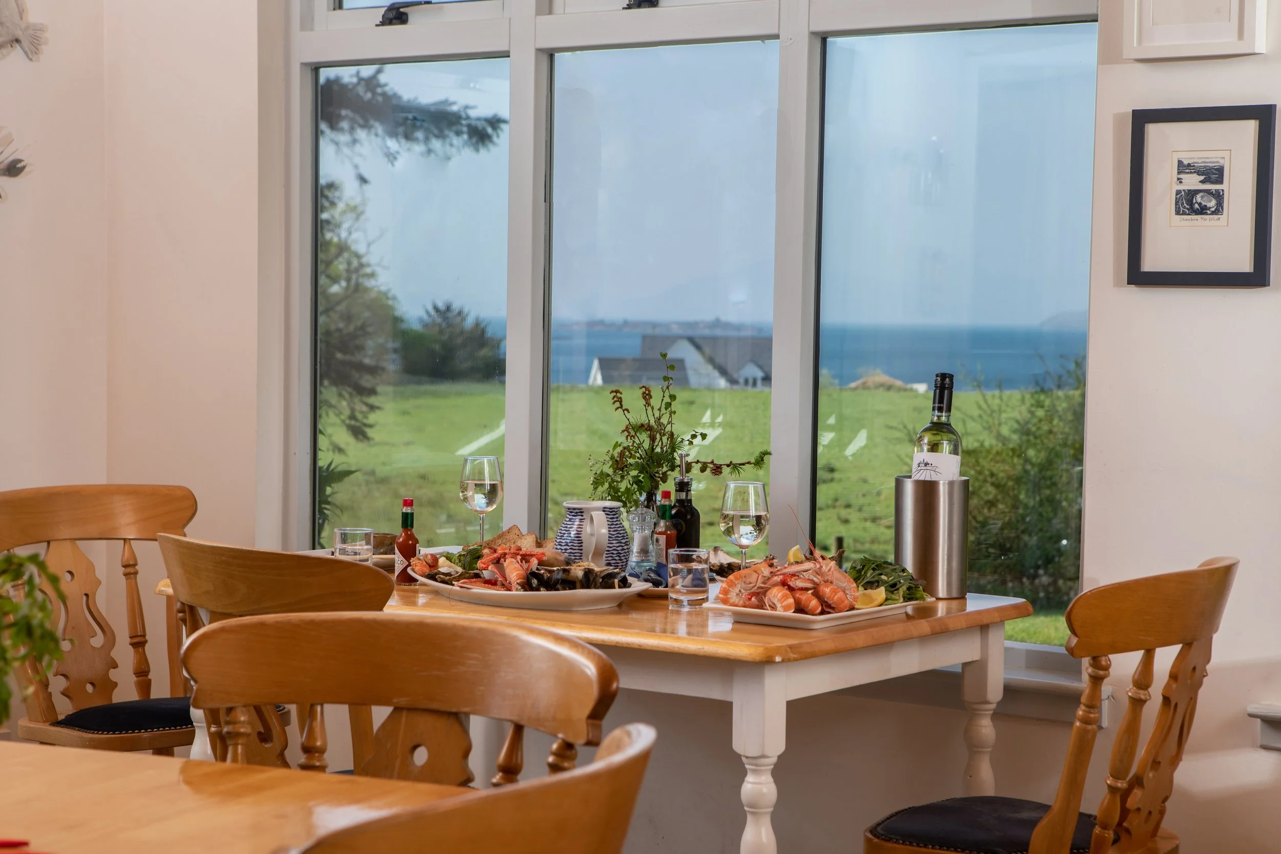 A dining room with a rectangular table set with seafood, drinks, and condiments, positioned near a large window overlooking a grassy landscape and water. Wooden chairs surround the table, and framed artwork decorates the wall.