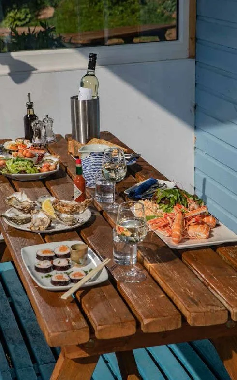 A wooden outdoor table set with various dishes including oysters on the half shell, sushi rolls, grilled vegetables, a salad, a bottle of white wine, glasses of water and wine, sauces, and beer bottles. The scene is illuminated by natural light through a window with a blue wall in the background.