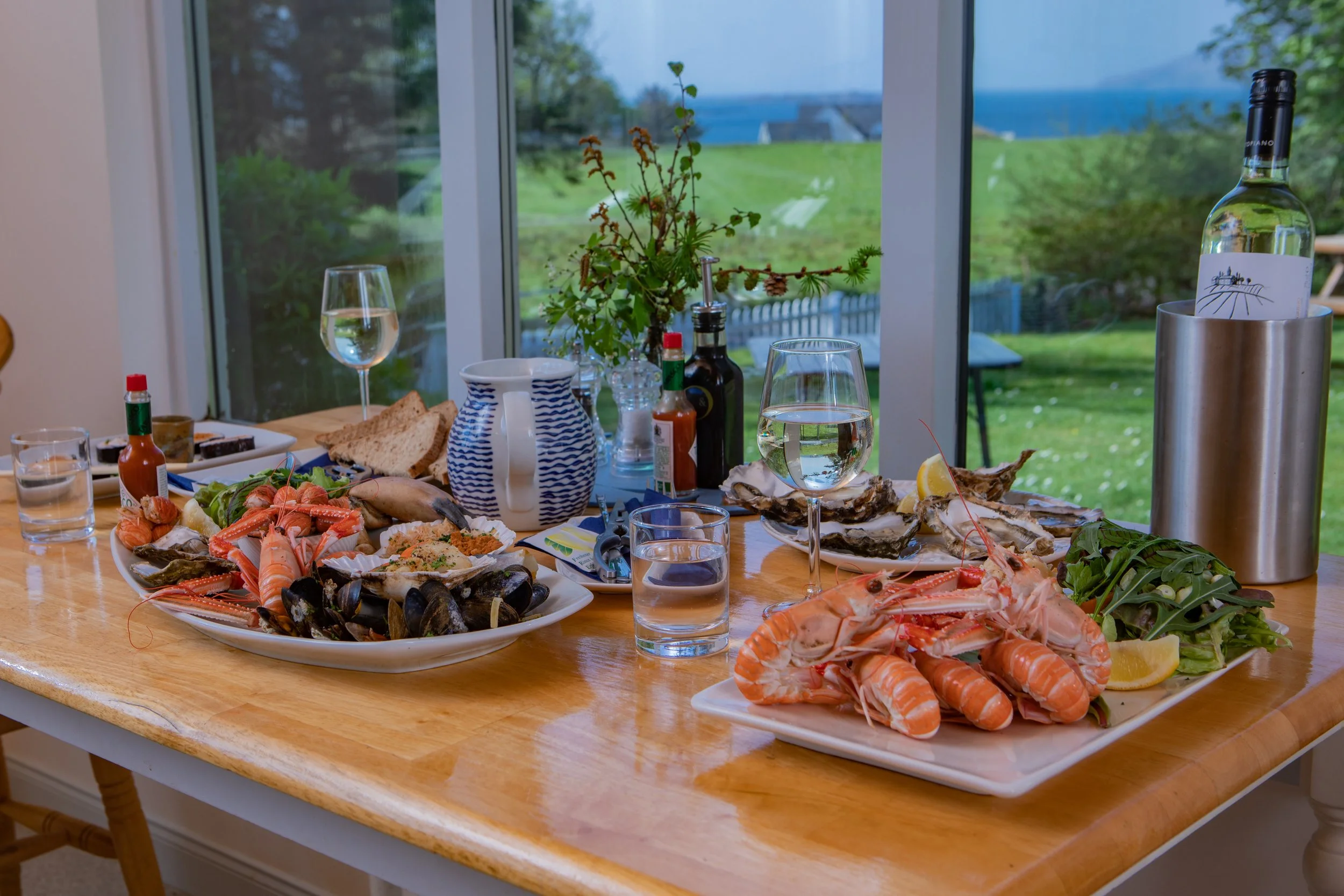 A dining table with seafood dishes, including lobster, shrimp, oysters, and various spreads, along with glasses of white wine and condiments. In the background, there is a window showing a lush green outdoor landscape.