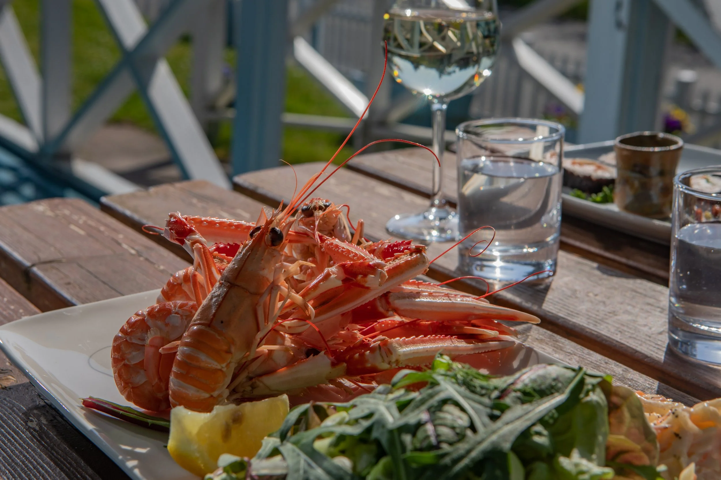 A plate of cooked lobster and seafood with lemon wedges, served on a wooden outdoor table alongside glasses of water and a salad.