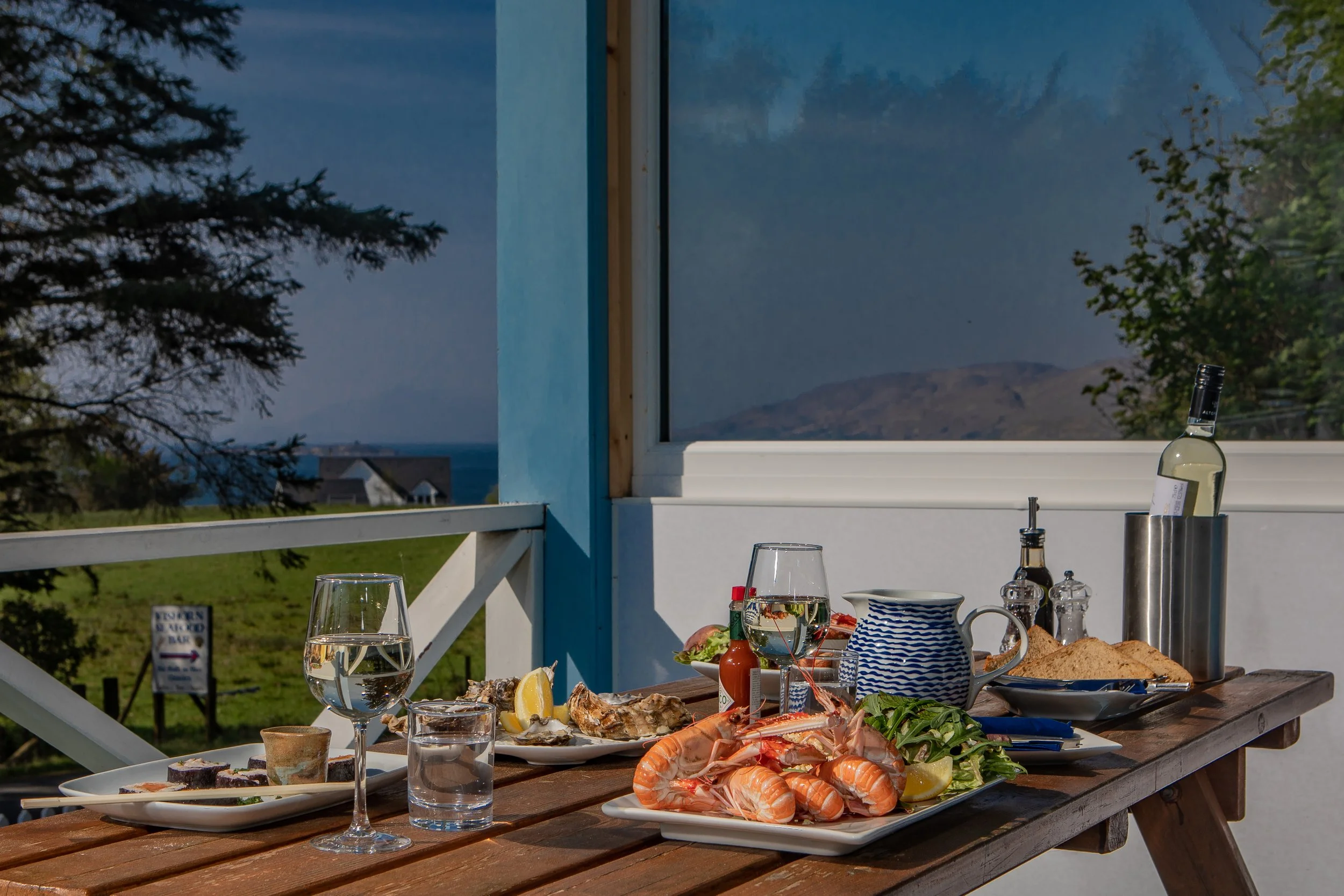 An outdoor dining table set with seafood, vegetables, and drinks on a wooden table, overlooking scenic hills and a blue sky through a large window.