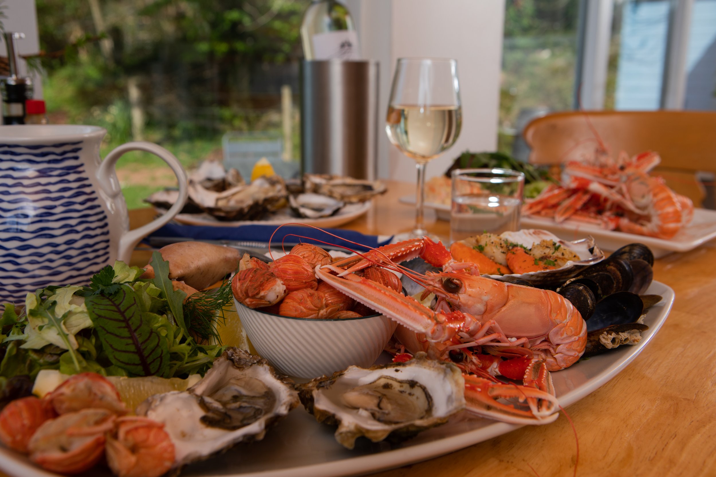 A table of seafood including oysters, shrimp, and lobster, with a glass of white wine and greenery in the background.