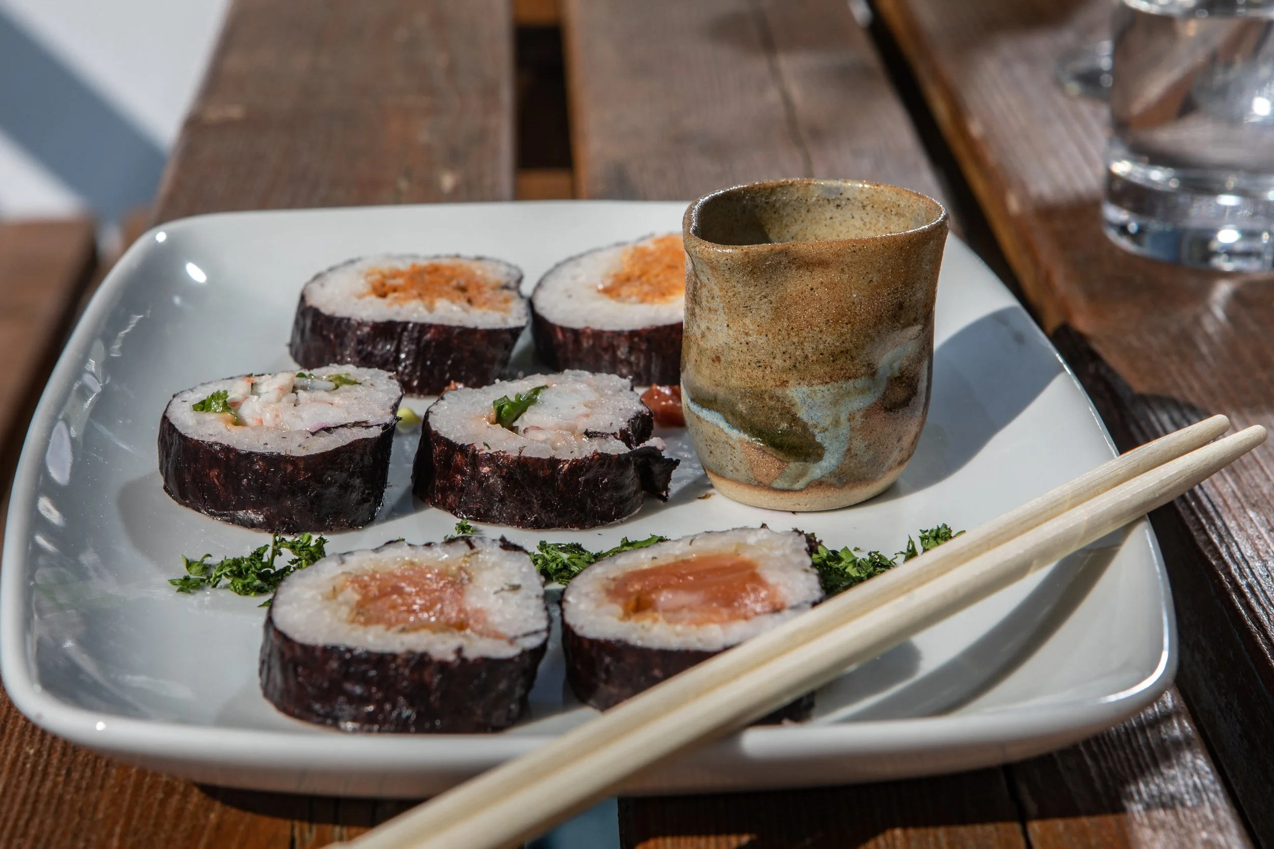 Sushi rolls on a white rectangular plate, with a ceramic cup and chopsticks, placed on a wooden table.