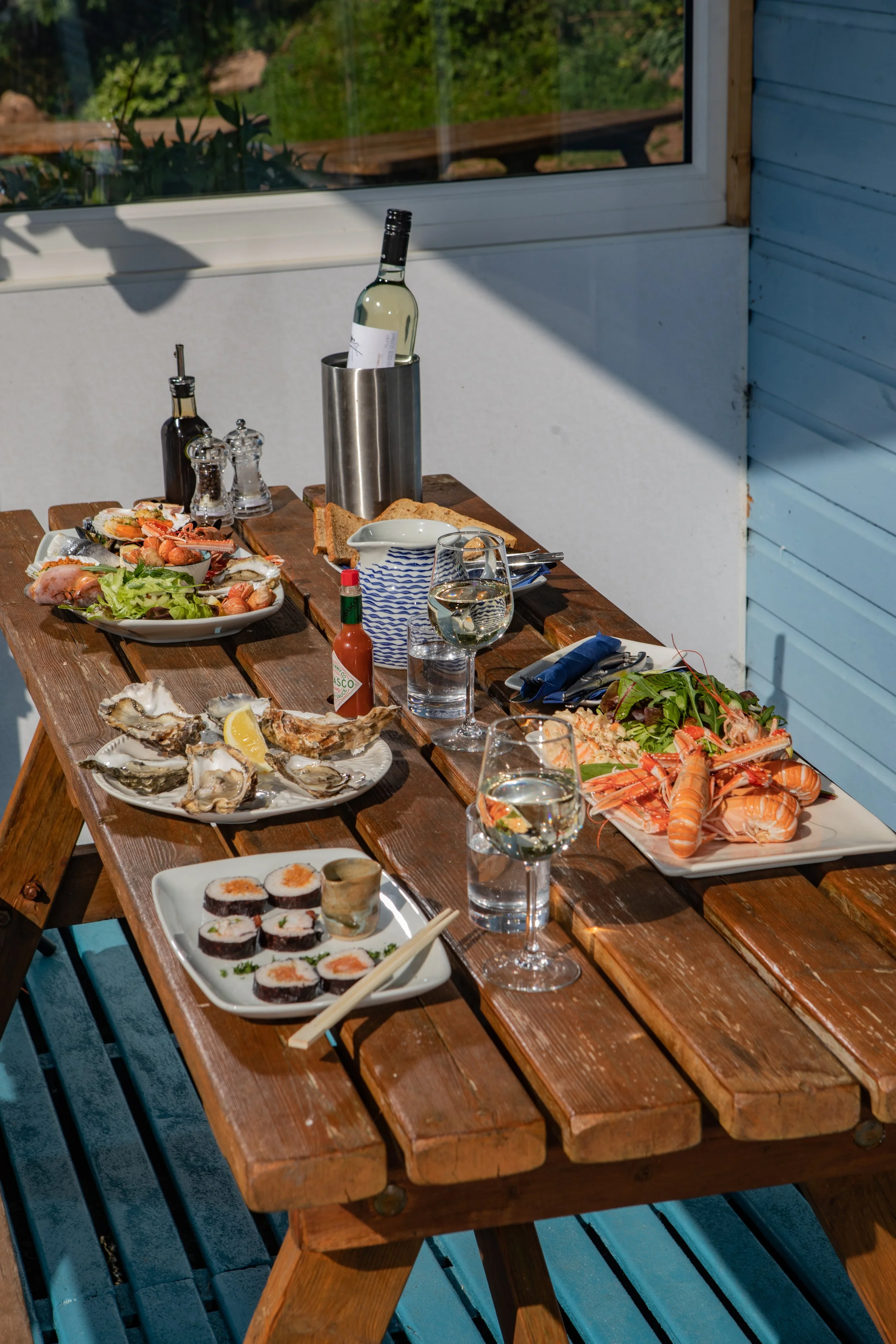 A wooden outdoor table filled with seafood and sushi dishes, including oysters, lobster, and sushi rolls, along with a glass of white wine, wine bottle in a stainless steel wine cooler, condiments, and a set of utensils, with a window and greenery vi