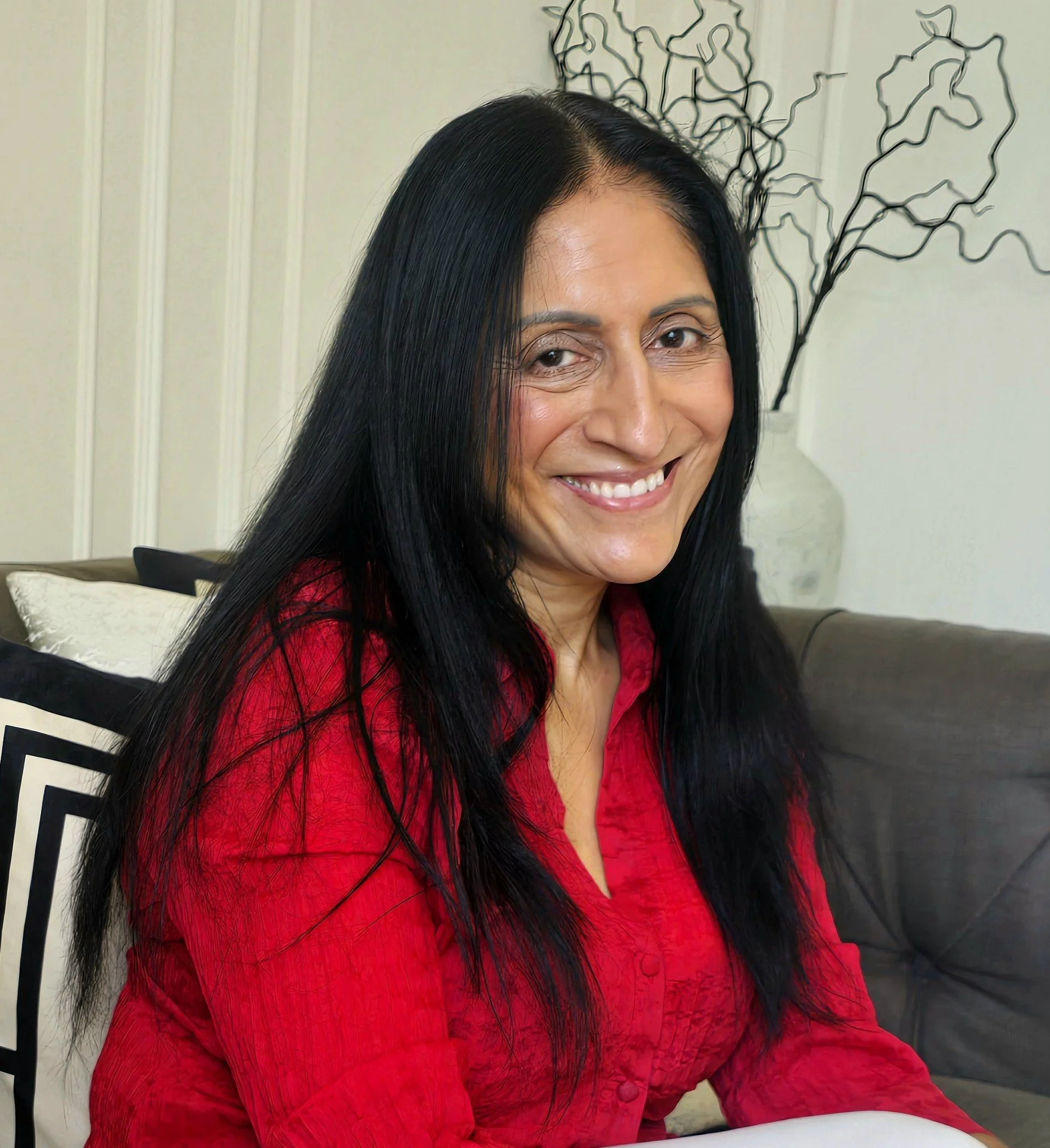 A woman with long black hair smiling, wearing a red textured shirt, sitting on a chair with a black and white patterned pillow, and a decorative black wire art piece in the background.
