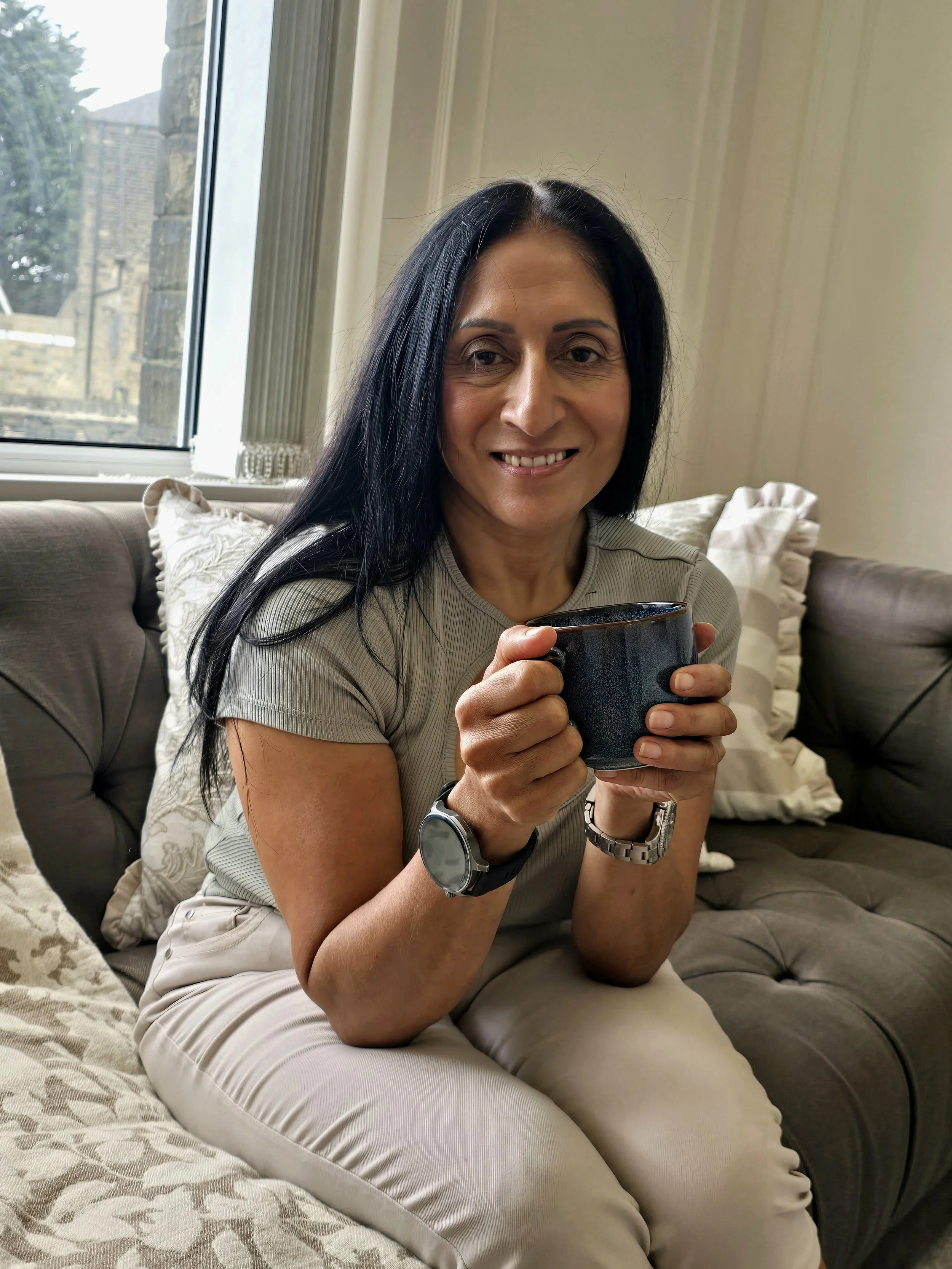 The author of Somebody Else's Life, Khatijah Shafi, sitting on a gray sofa, holding a dark mug leaning forward, smiling, in a cozy room with a window showing an outside view.