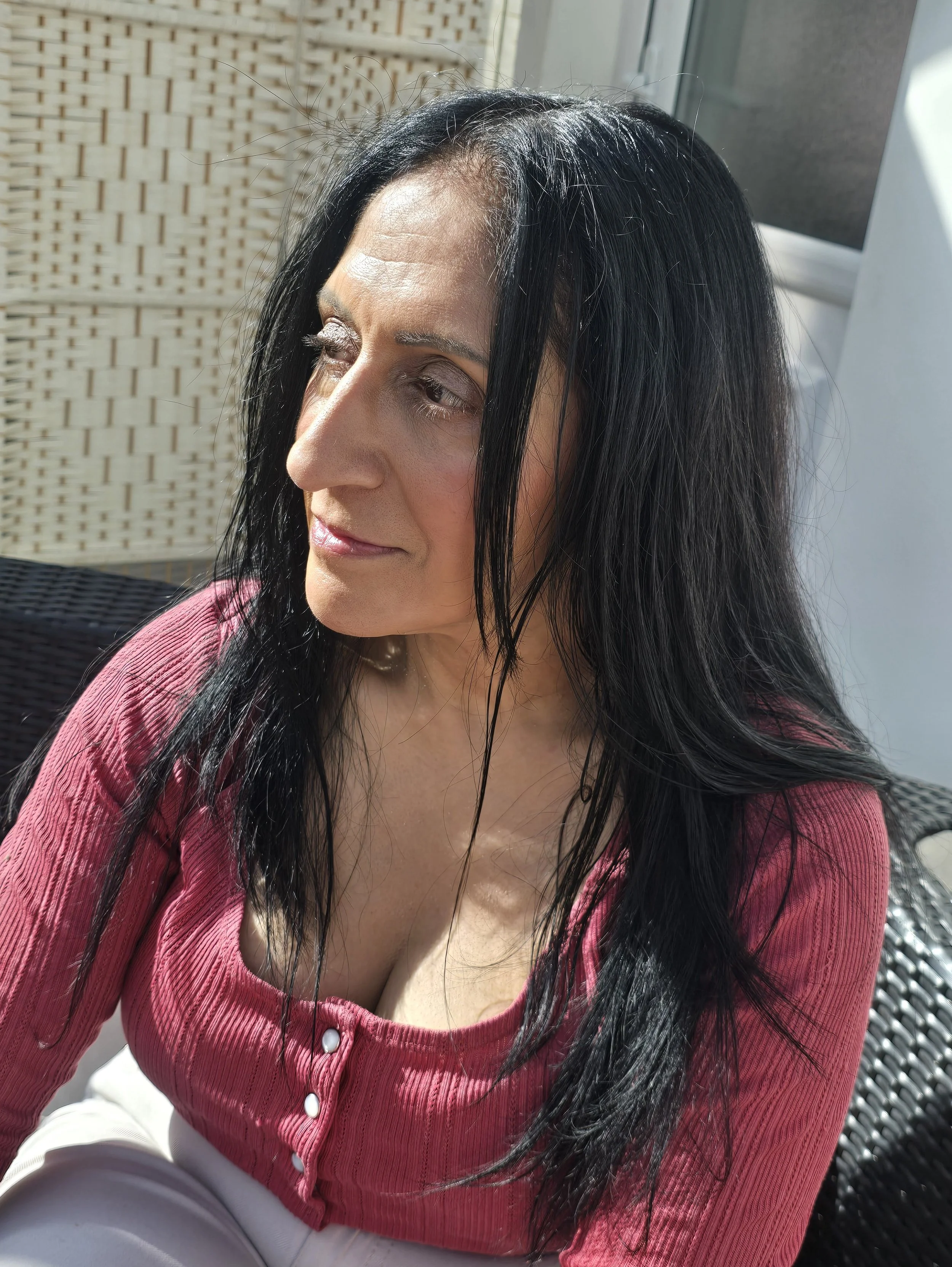 Close-up of a woman with long black hair wearing a pink sweater with white buttons, sitting outdoors in sunlight.