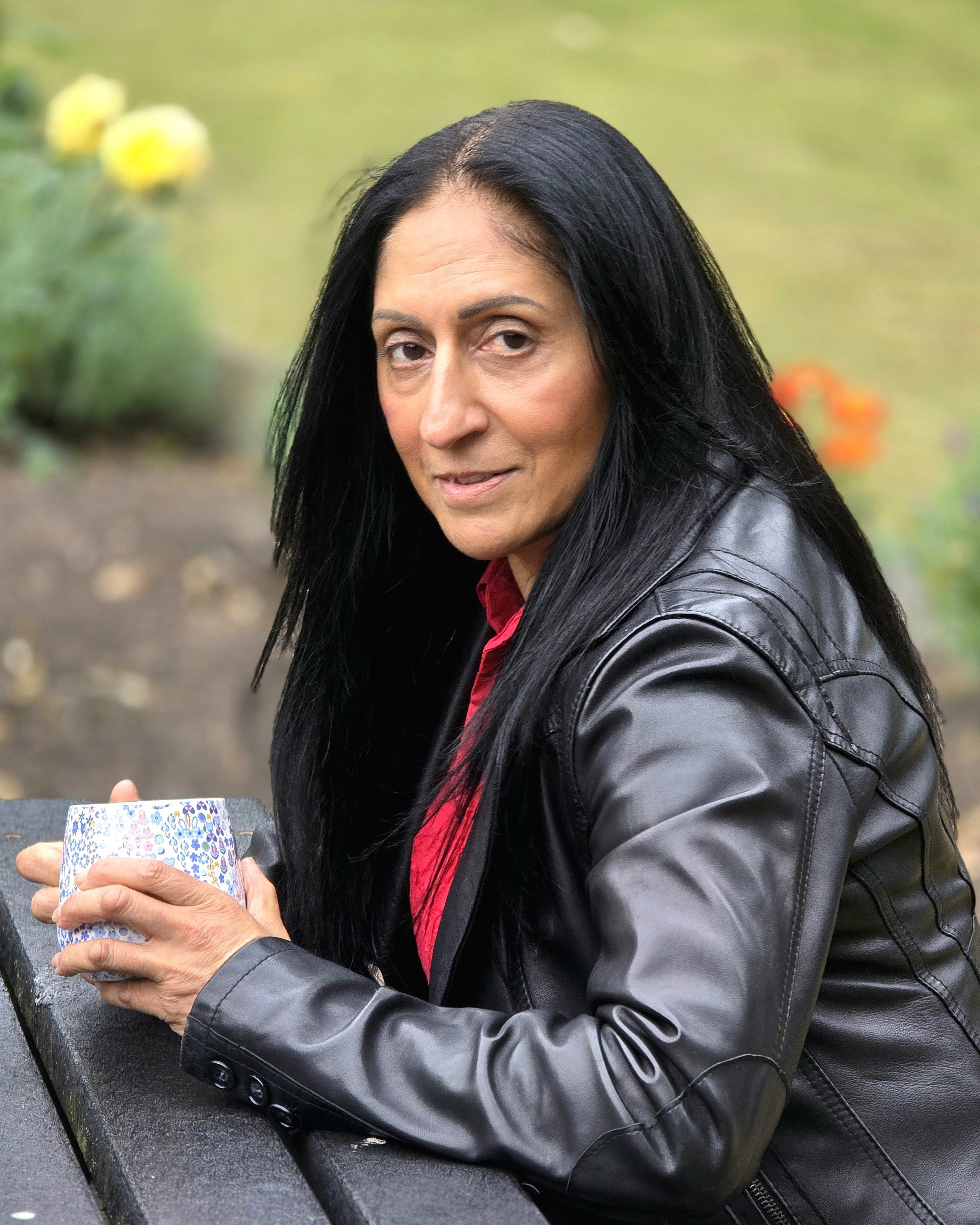 A woman with black hair wearing a leather jacket and red shirt, holding a colorful cup with her left hand, sitting on a park bench.