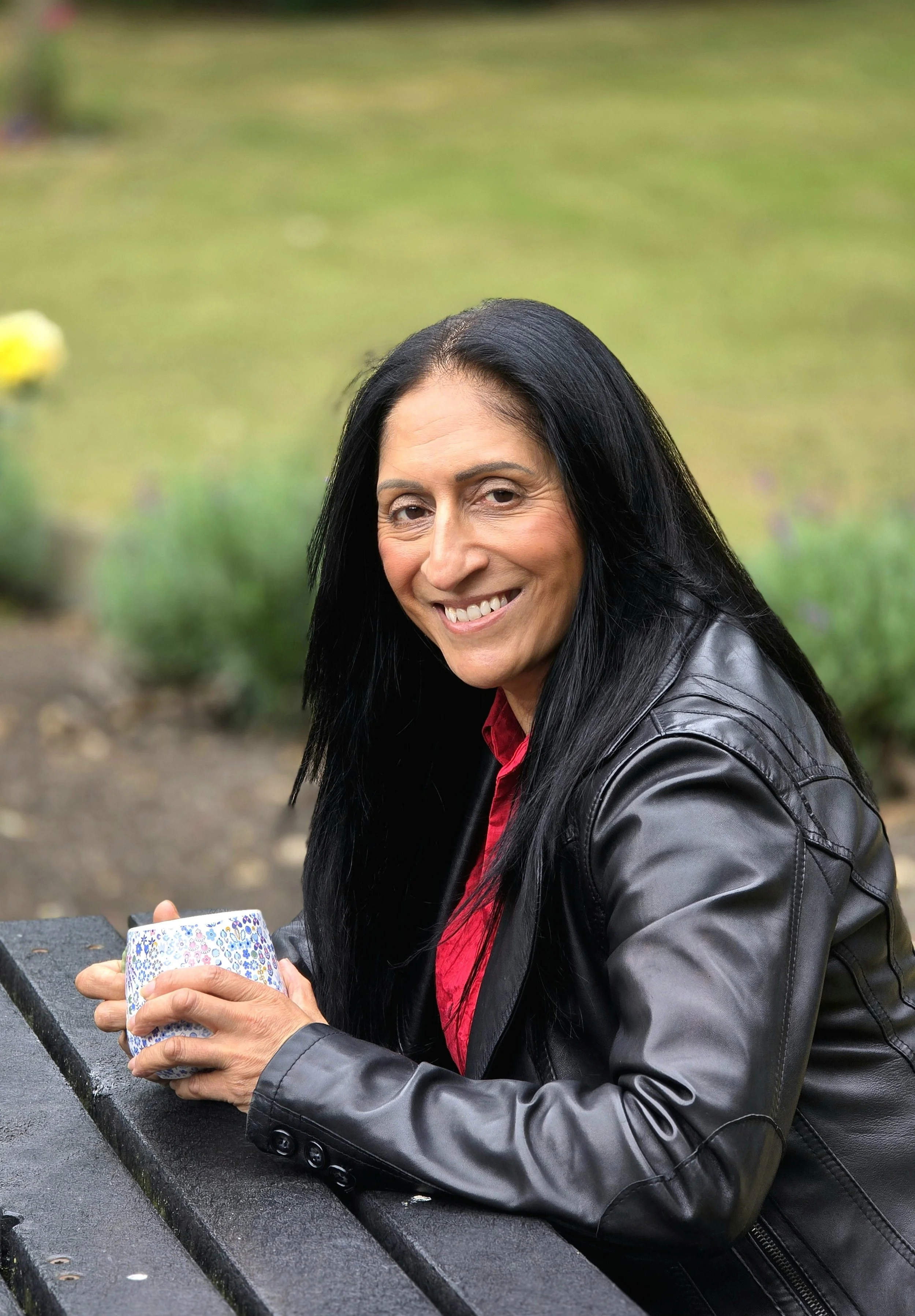 The author of Somebody Else's Life, Khatijah Shafi, wearing a black leather jacket sitting at a bench outdoors, holding a colorful patterned mug, smiling at the camera.