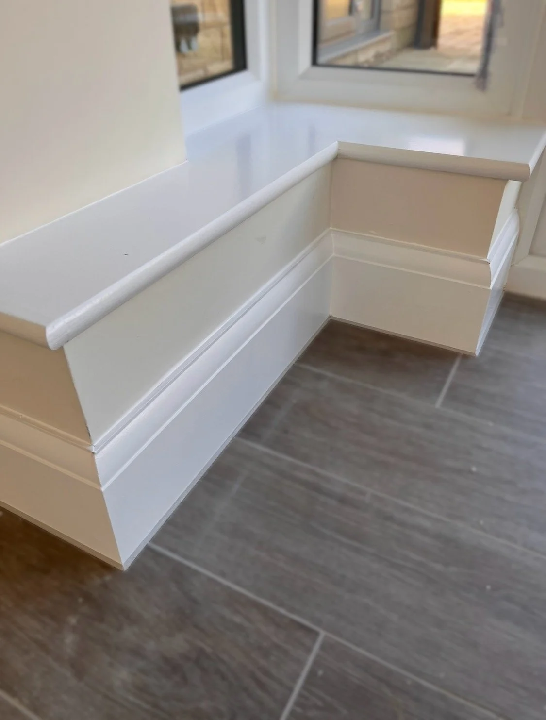 Close-up of a white built-in window seat or bench with a ledge, next to a window with a view outside, and a brown tiled floor.