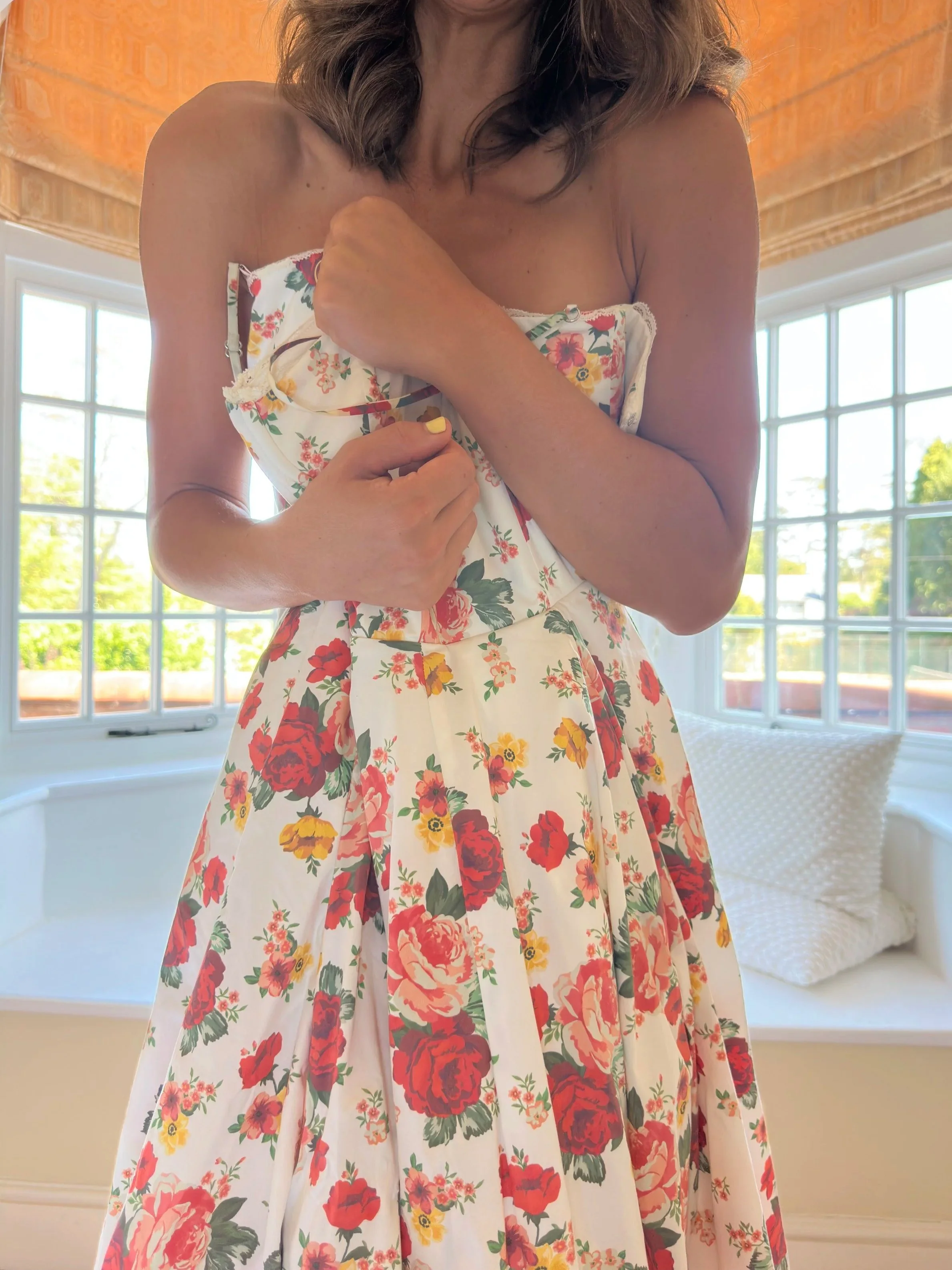 Woman holding an unzipped floral dress with red, yellow, pink, and green flowers, close to her chest inside a room with large windows and partly drawn blinds.