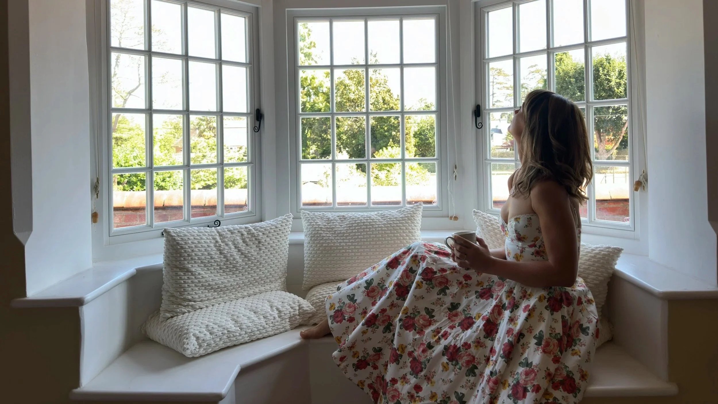 A woman in a floral dress sitting on a window bench holding a mug, with large grid-patterned windows showing green trees outside, and plush pillows on the bench.