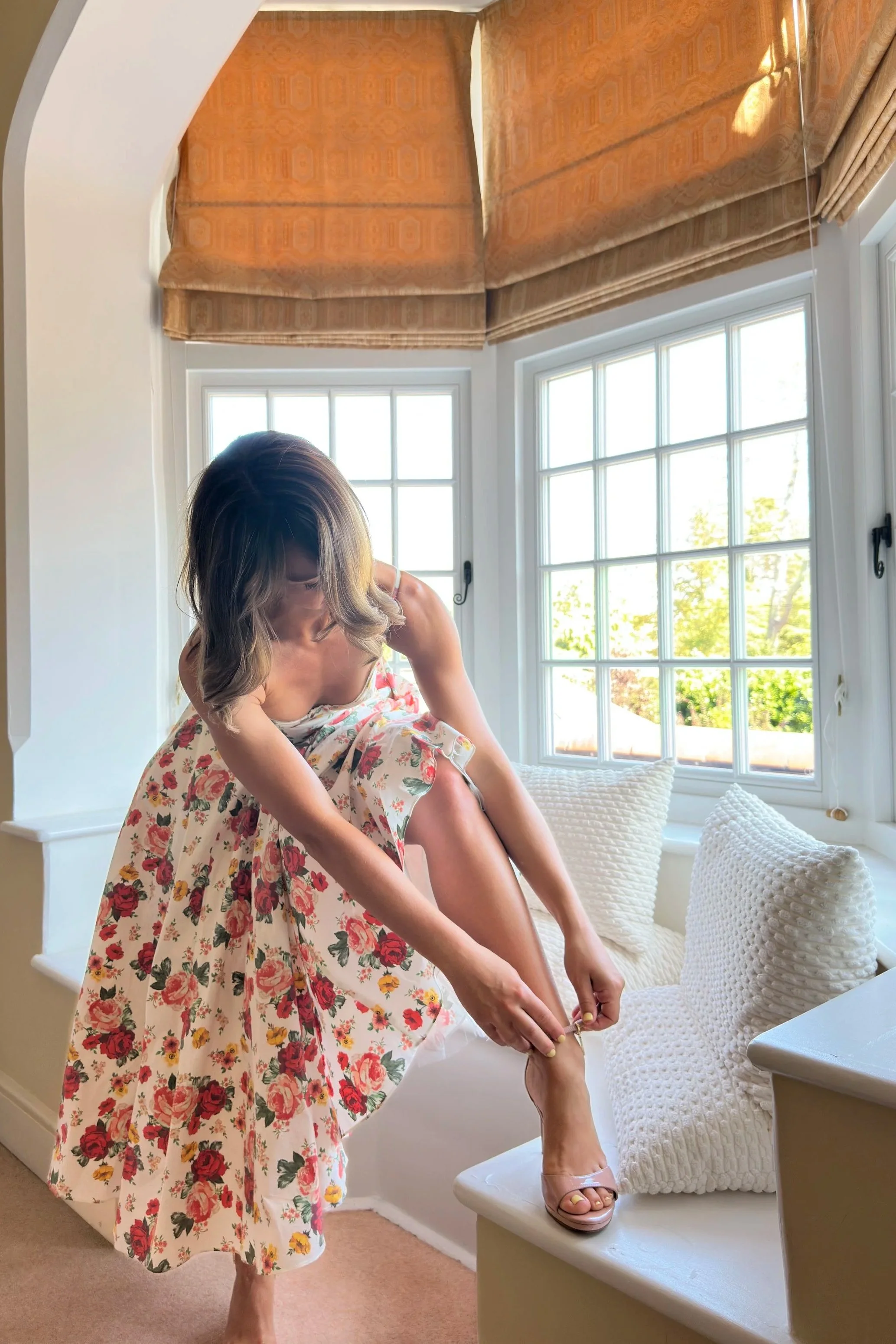 A woman in a floral dress is putting on a pink sandal on her right foot on a window seat. The window behind her has white framing and is partially covered by tan curtains. The window seat has cream textured pillows and a white knitted blanket, with a small white table beside it. The room has a light-colored carpet and beige walls.