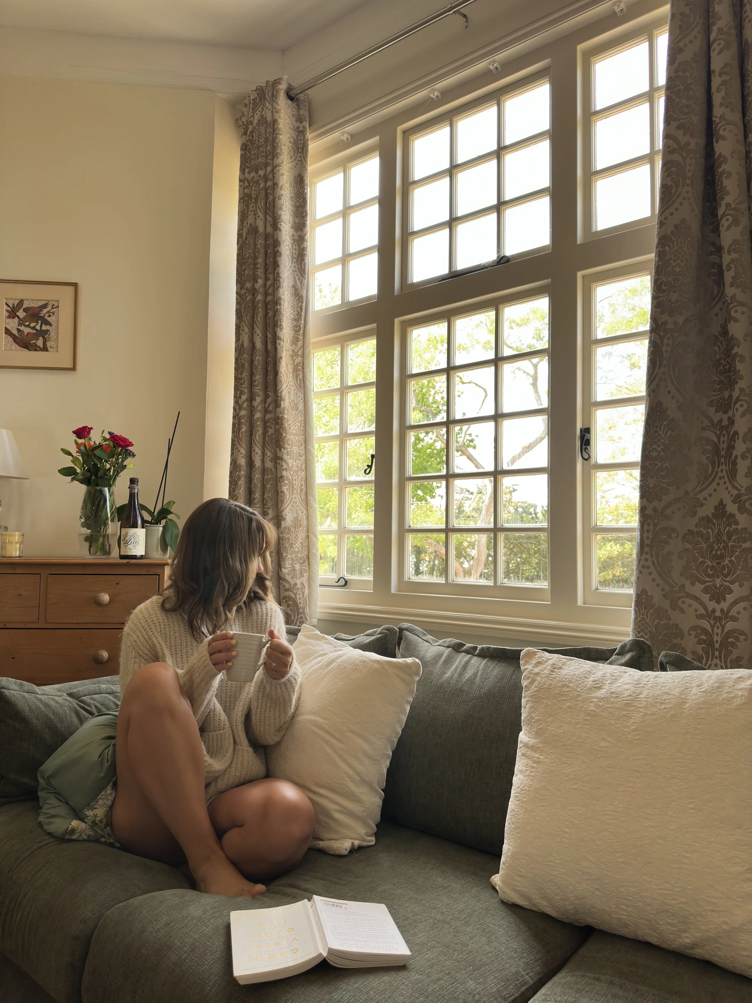 A brunette woman in a sitting room with large windows, holding a white mug, with an open book on the sofa in front of her. The room is decorated with beige curtains, a wooden dresser with a vase of pink and red flowers.