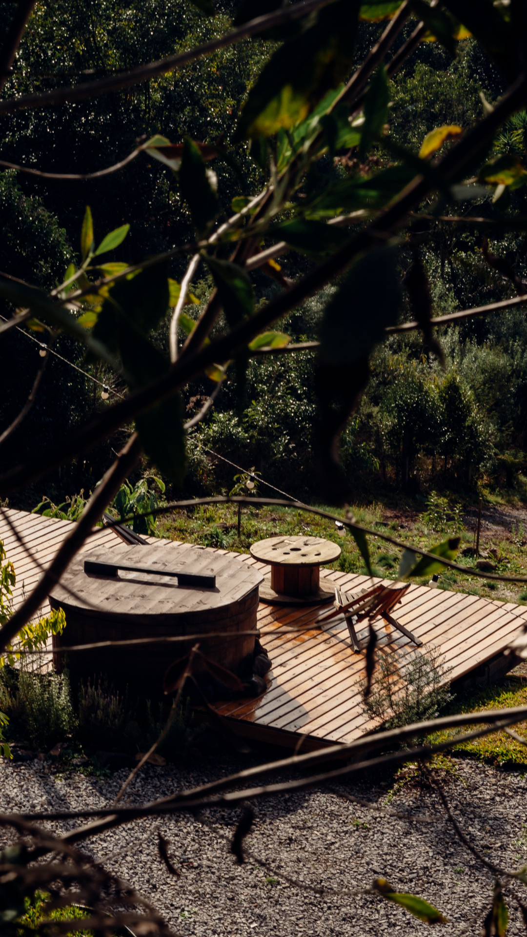 A wooden deck with a round wooden table and a lounge chair, surrounded by trees and greenery.