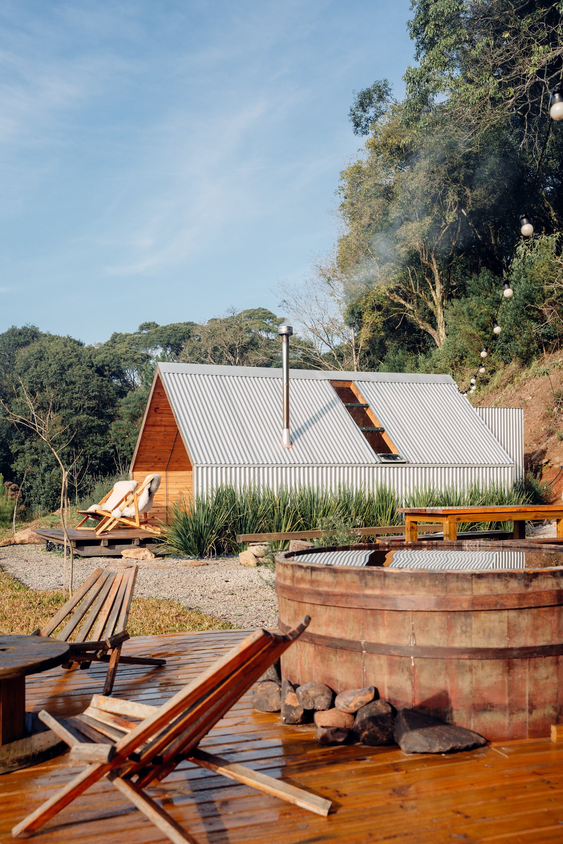 Exterior view of a small modern cabin with a metal roof, surrounded by trees and outdoor furniture, including lounge chairs and a large fire pit, under a clear blue sky.
