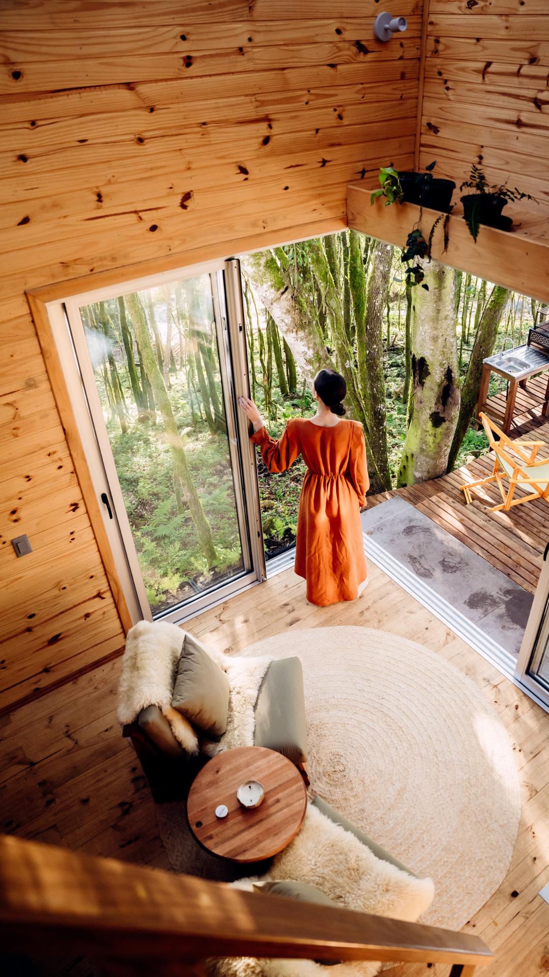 A woman in an orange dress stands by a large glass sliding door, looking out into a lush green forest. The interior features wooden walls and flooring, with a cozy beige and cream-colored armchair with pillows, a round beige rug, and a small wooden side table with candles. Outside, there is a wooden deck with chairs and a small outdoor sink.