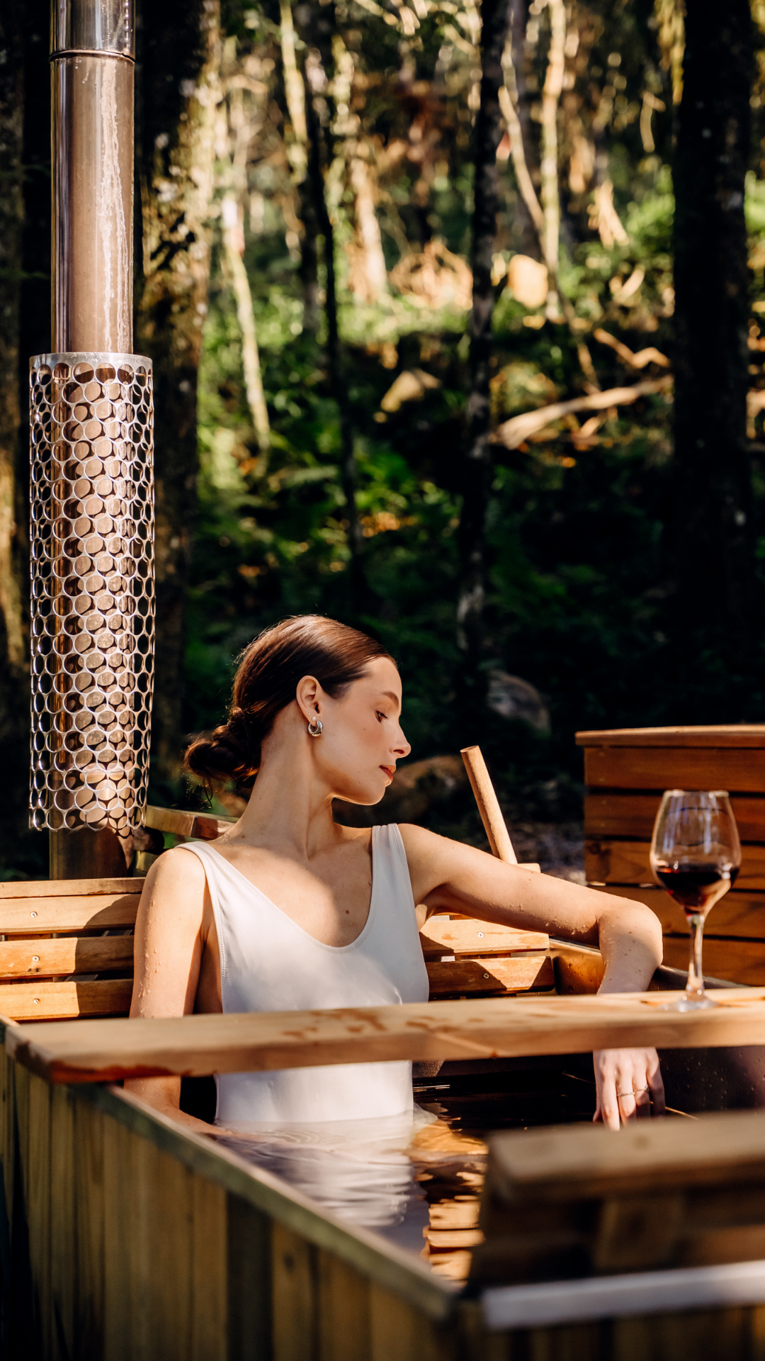 Woman wearing a white sleeveless top relaxing in an outdoor wooden hot tub with a glass of red wine on the side, surrounded by a forest.