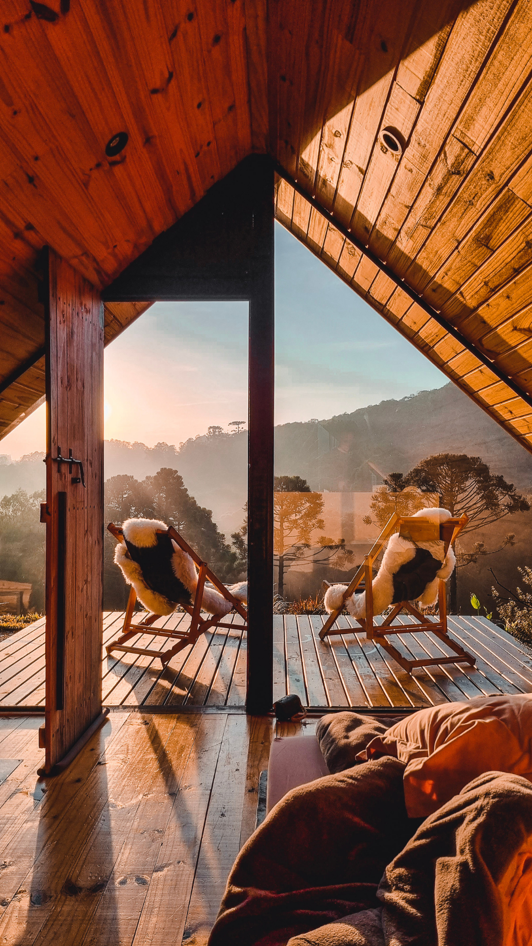 View from a wooden porch with two lounge chairs covered in sheepskin, overlooking a misty sunset landscape with trees and hills.