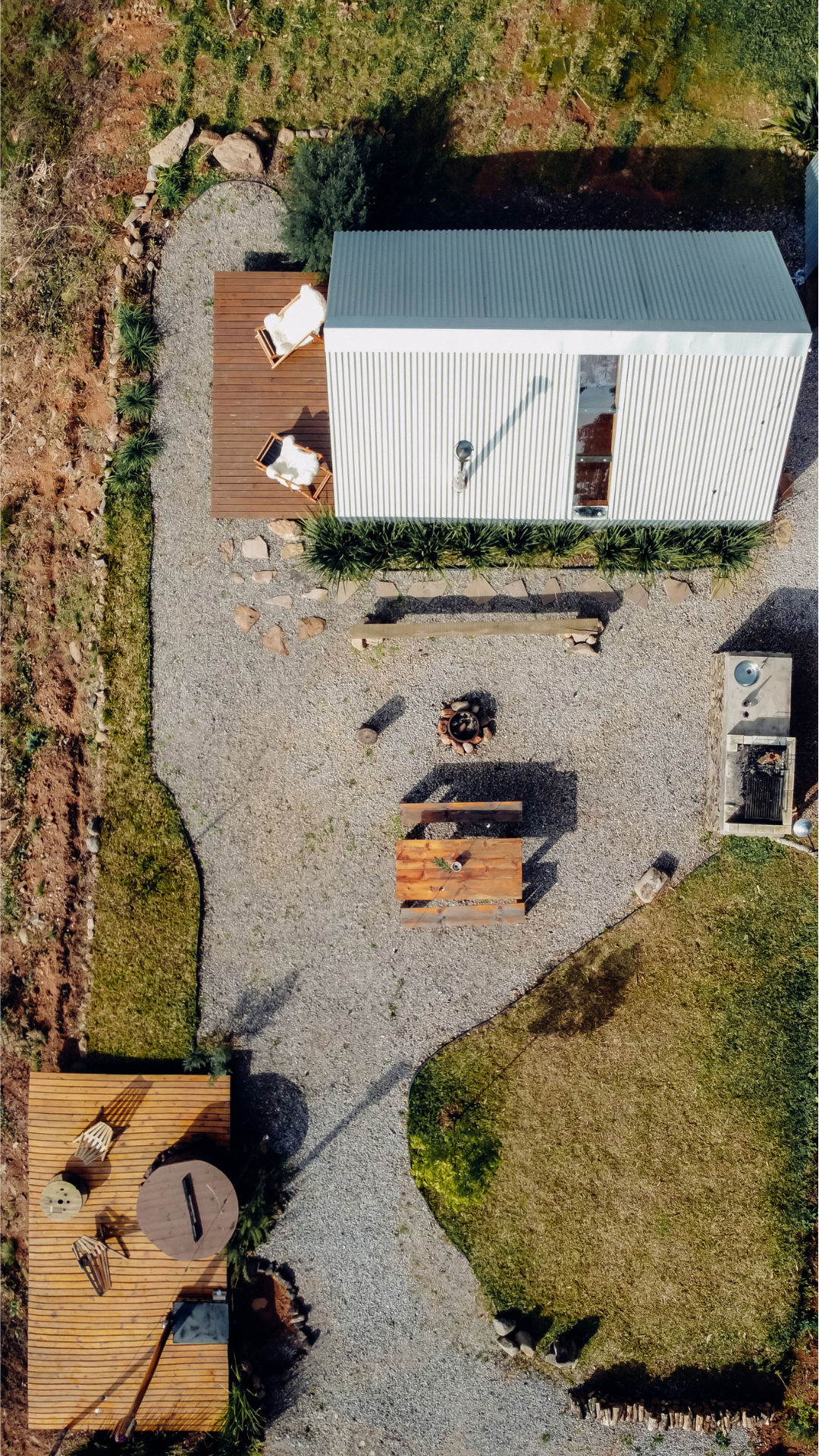 Top-down aerial view of a backyard with a gravel pathway, a small wooden deck with outdoor chairs and a table, a fire pit, a barbecue grill, and a modern metal-roof shed with outdoor seating.