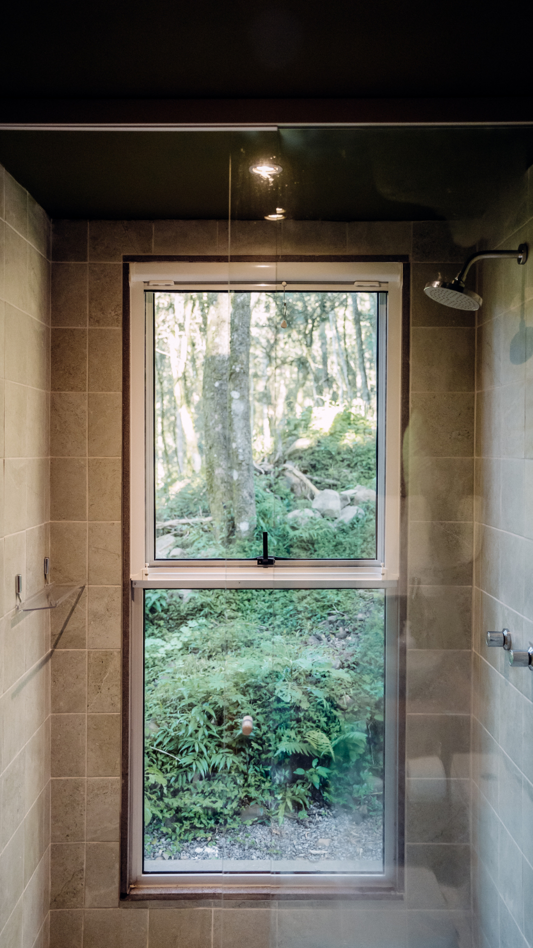 A shower with beige tile walls and a window overlooking a green forest.