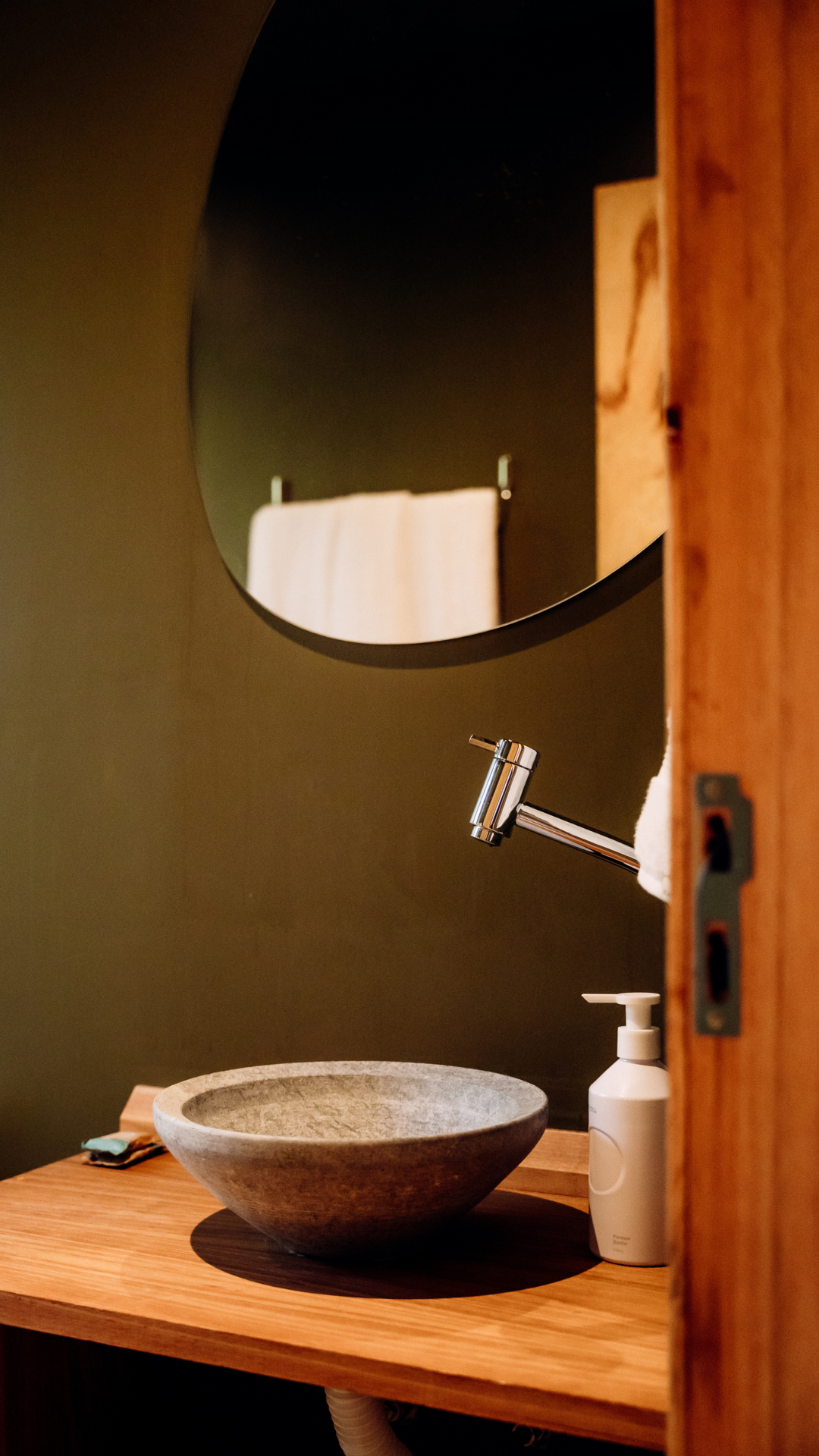 A bathroom with a round mirror, a metallic faucet, a stone sink, a soap dispenser, and a towel on a wooden countertop.