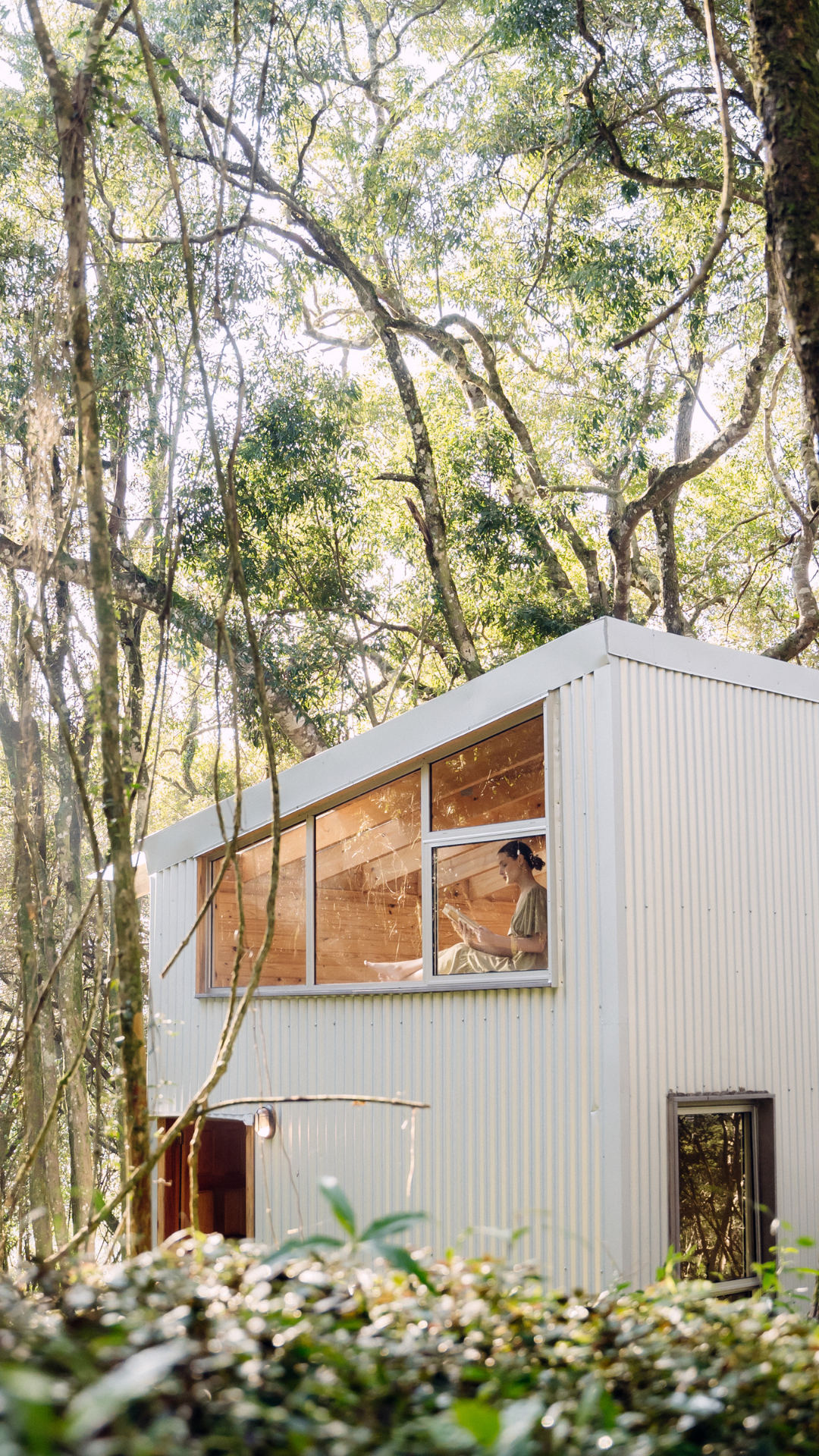 A woman sitting inside a modern, white, two-story house with large windows, reading a book while surrounded by trees and greenery in a forest.