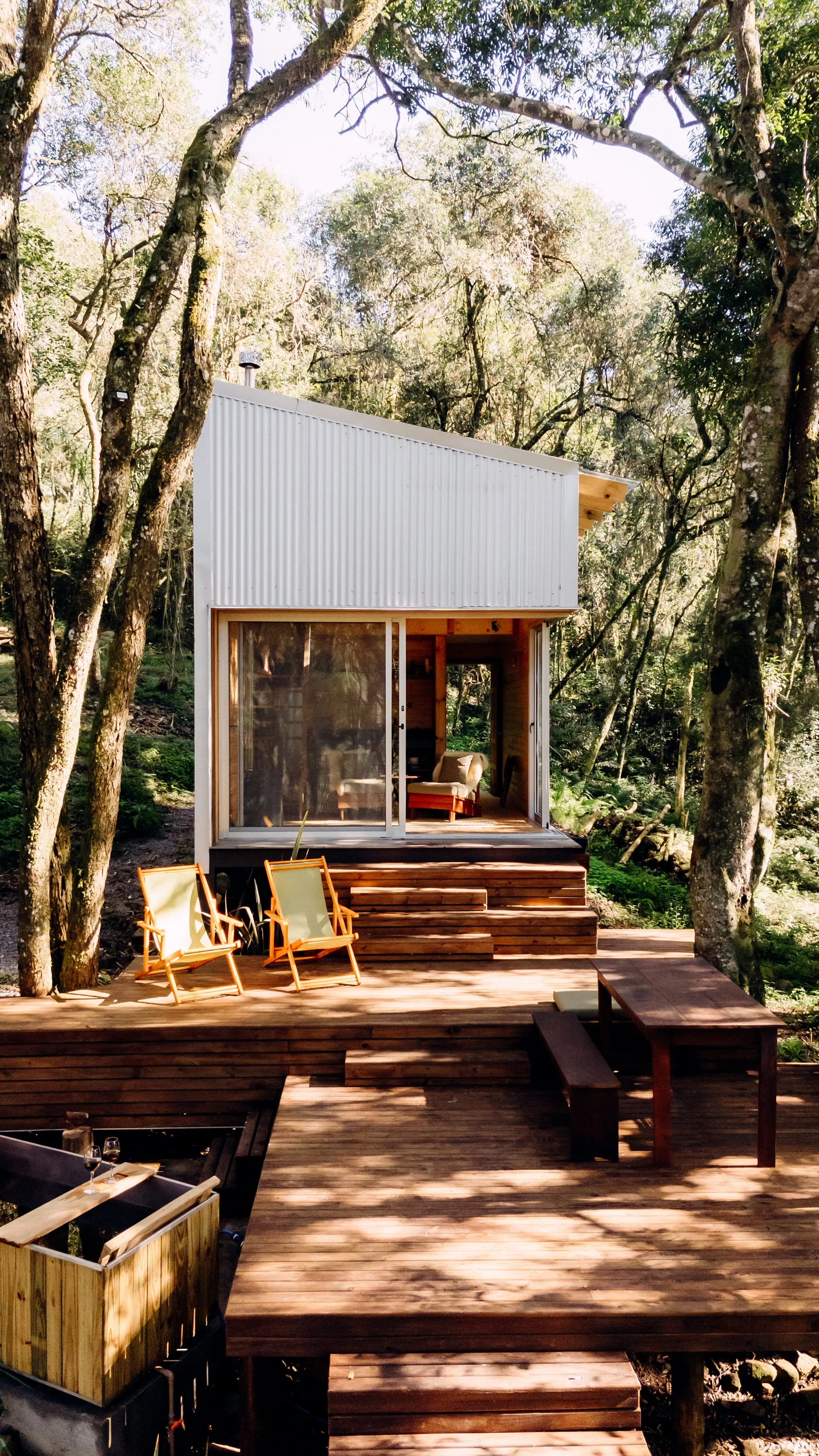 A modern tiny house with white metal siding and large glass sliding doors, situated in a wooded area with trees surrounding it. There is a wooden deck with steps leading up to the house, outdoor seating including two orange chairs and a wooden bench, and a small table. The interior of the tiny house features a cozy living space with a couch visible through the glass doors.