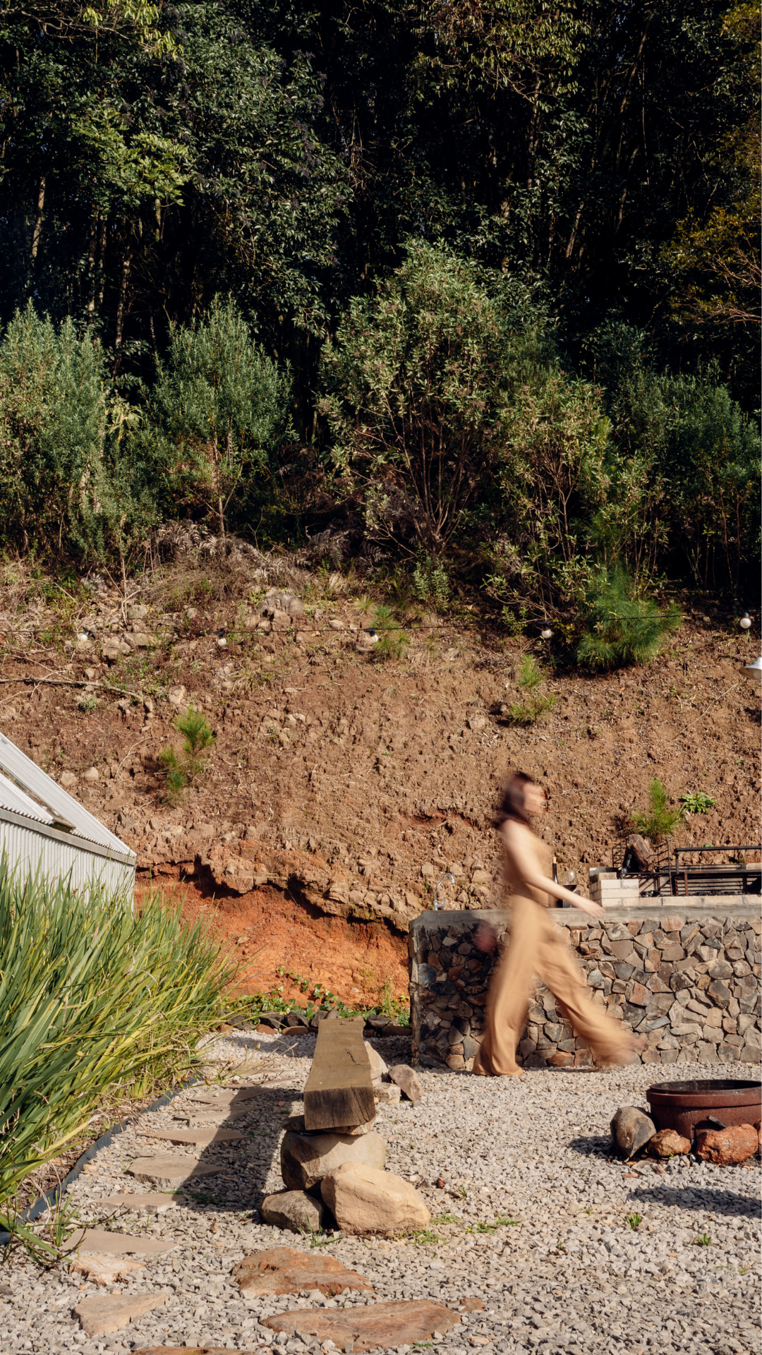 A woman walking along a stone and gravel path, with a sloped hillside covered in greenery and small shrubs behind her. There are outdoor elements such as rocks, a wooden log, and a stone wall.